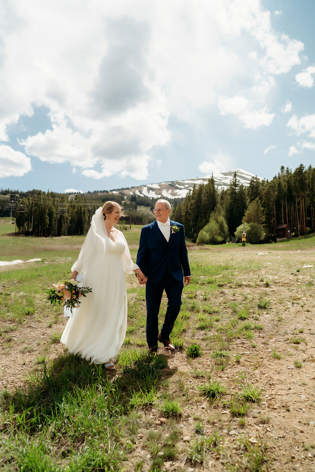 Barefoot and smiling, the bride and groom walk through an open meadow under a blue sky with ski slopes in the distance.