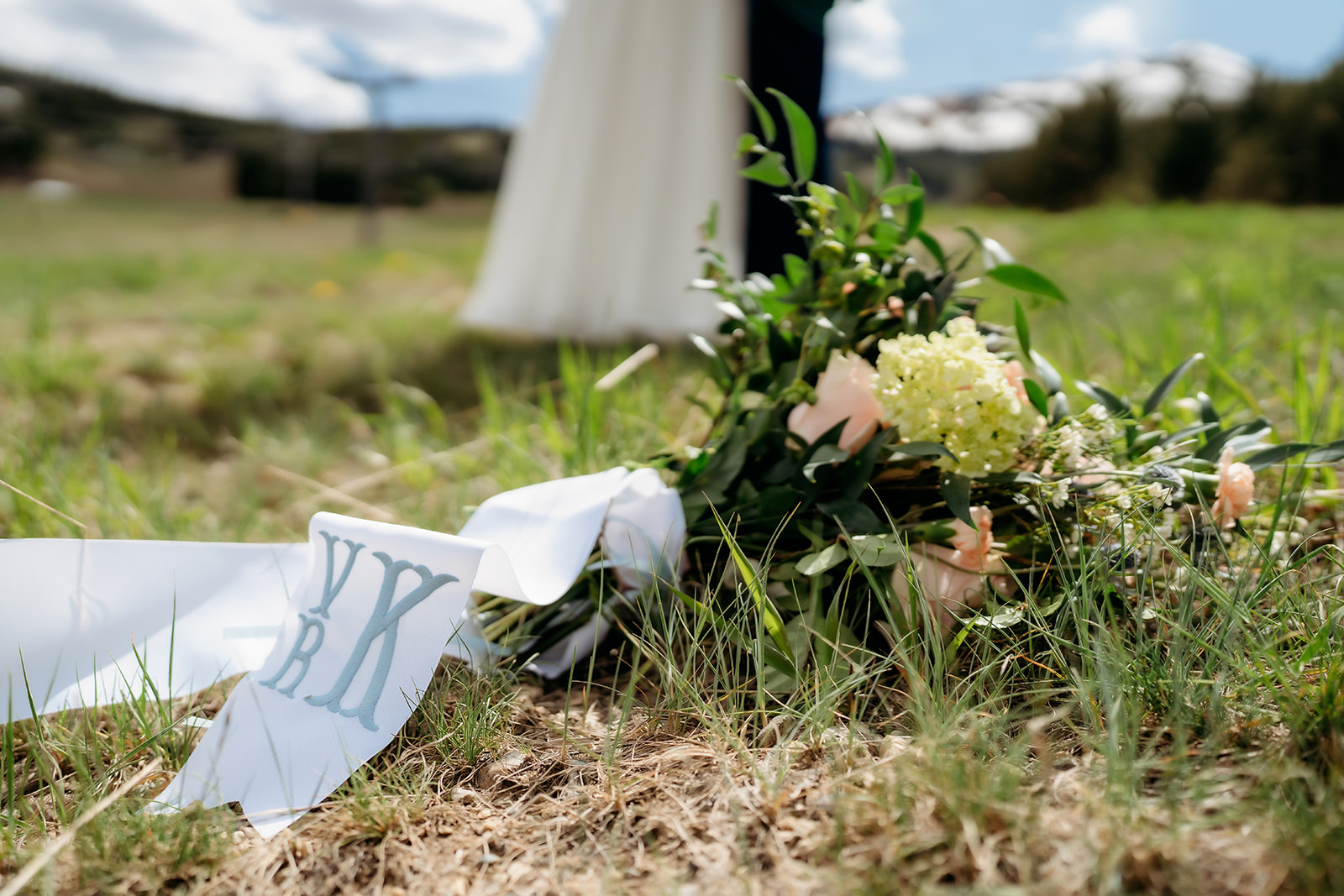 A lush bouquet of blush blooms and greenery lies in the grass, ribbon with custom monogram fluttering beside the bride’s flowing dress.