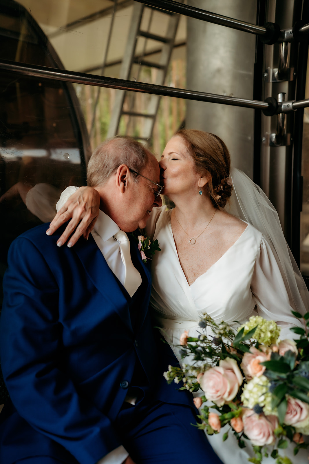 A soft, emotional moment as the bride gently kisses her groom’s forehead, surrounded by the warm glow of natural light and reflections.