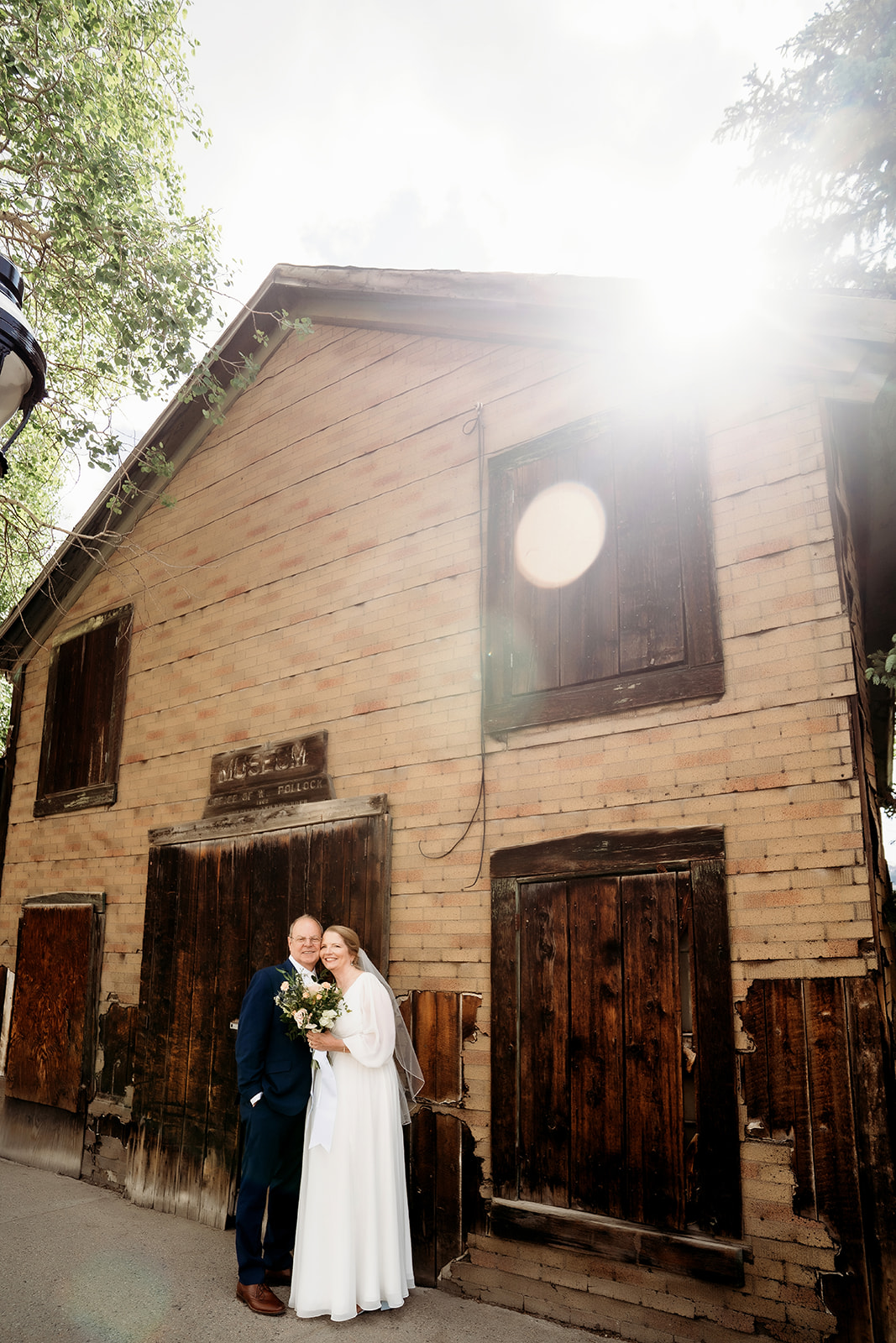 A soft sun flare shines behind the couple as they pose in front of an aged wooden building, blending history with romance.