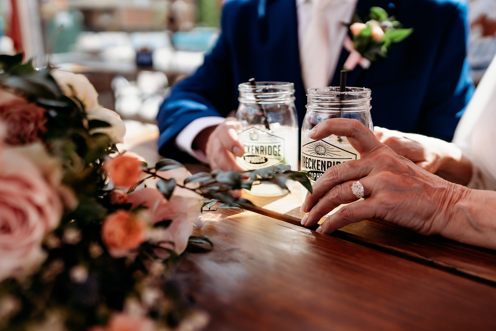 A close-up of the bride and groom’s hands as they enjoy cocktails in mason jars, surrounded by soft florals.