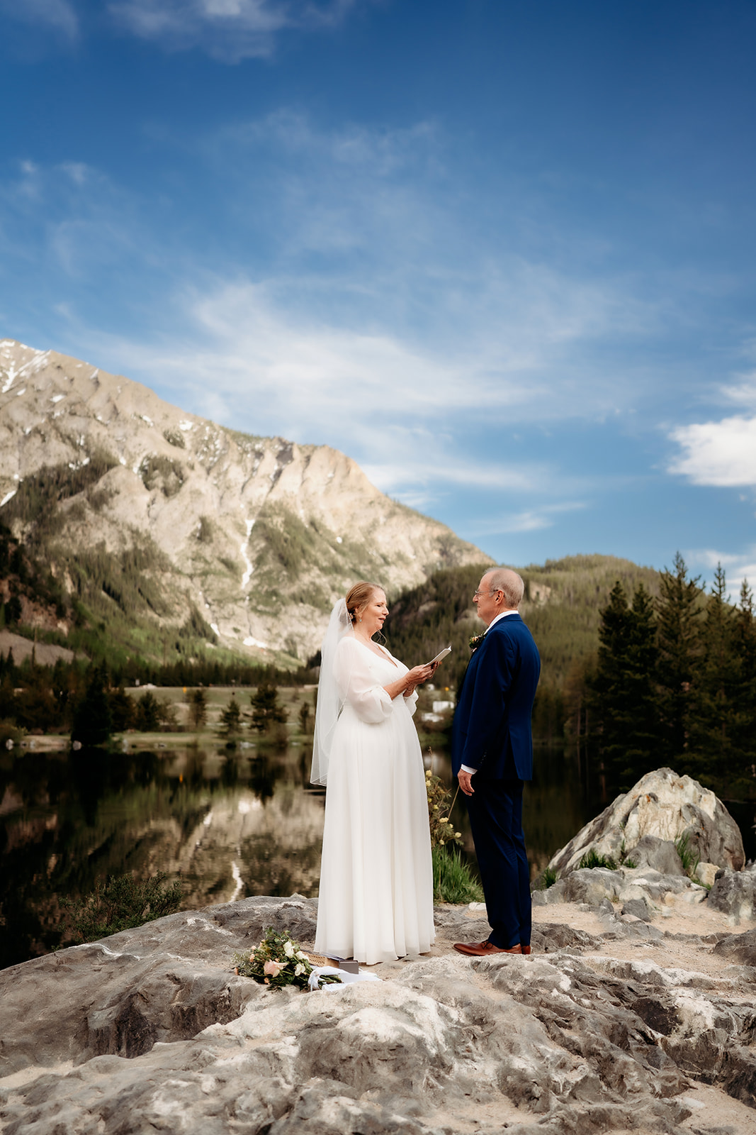 A bride and groom stand on a rocky overlook, exchanging vows with snow-capped peaks and blue sky behind them—one heartfelt way to show How to Make Your Elopement Special.