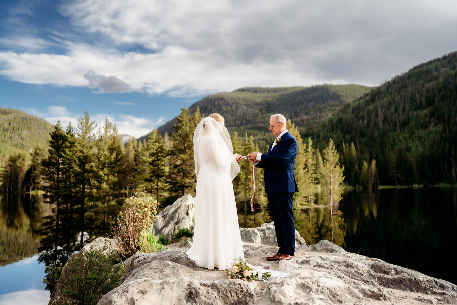 The couple shares a meaningful handfasting ritual atop a rock above a reflective alpine lake, surrounded by forest and mountains.