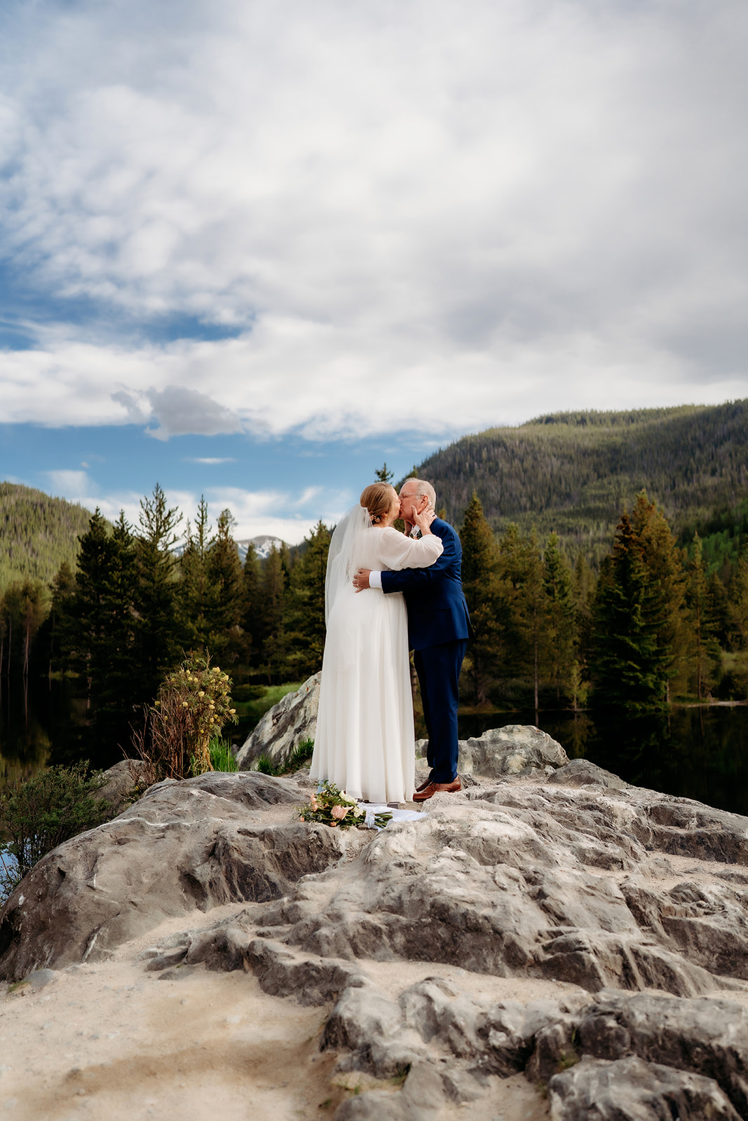 The couple wraps each other in a post-ceremony hug against a backdrop of green trees and a vibrant sky.