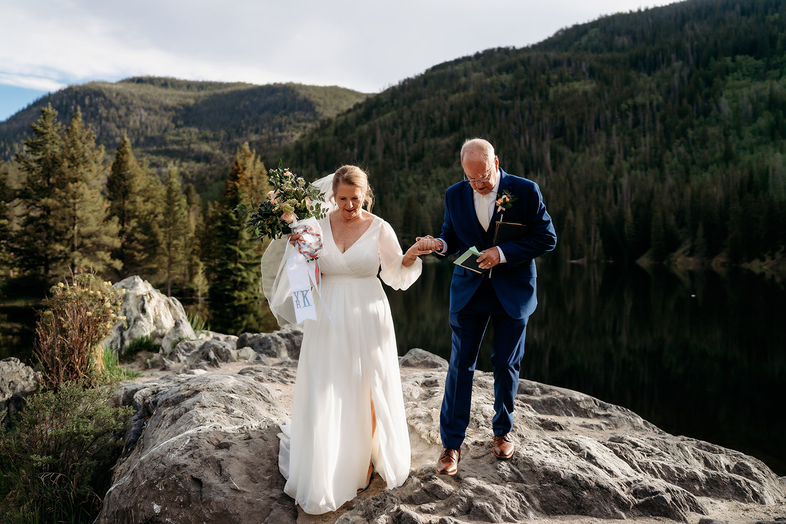The groom gently assists the bride as they walk down a rugged trail after their vows, bouquet in hand and love in every step.