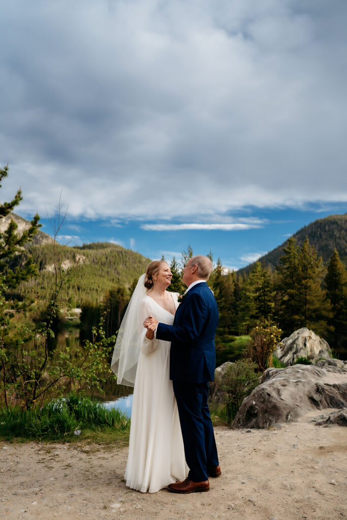 The couple dances together in front of a lake and pine-covered mountains under a dramatic blue sky.