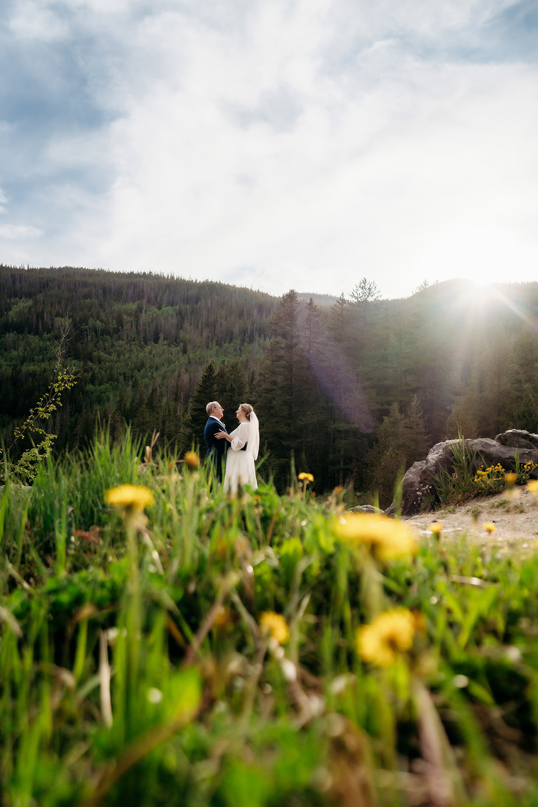 A cinematic photo of the couple holding hands in a wildflower field with mountains and sun flare in the background—capturing How to Make Your Elopement Special with nature’s beauty.