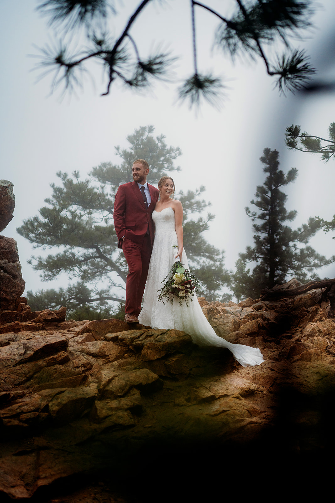 Bride and groom gazing out together, standing hand in hand on rugged rocks in the mist.