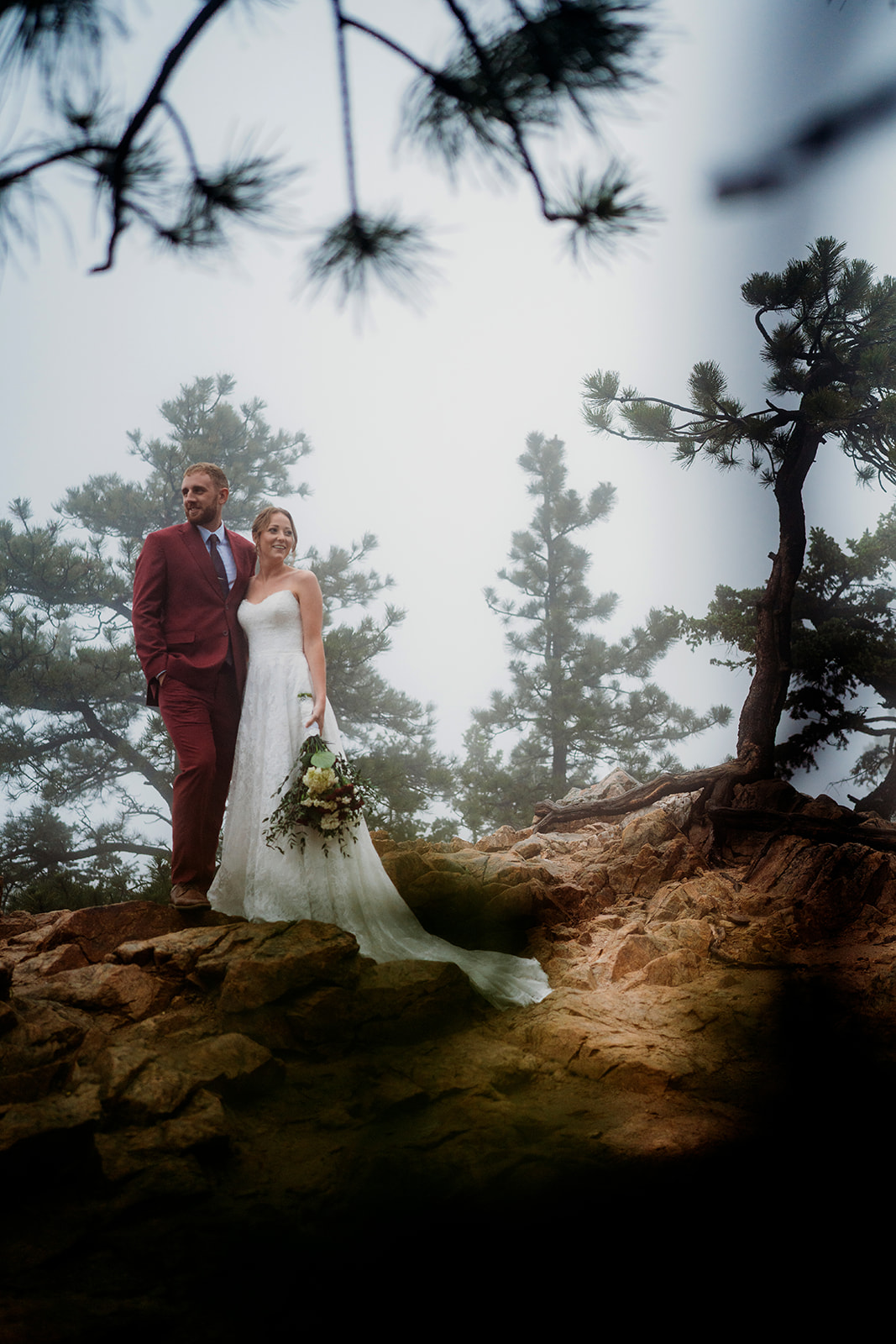 Bride and groom standing close together on a rocky outcrop, surrounded by pine trees.