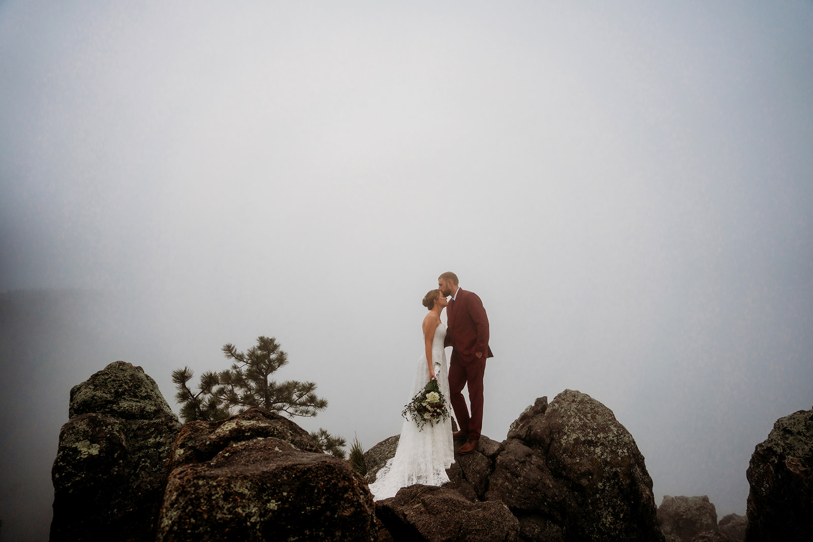 Couple sharing a kiss on rocky cliffs surrounded by fog, highlighting how to plan an elopement in the mountains.