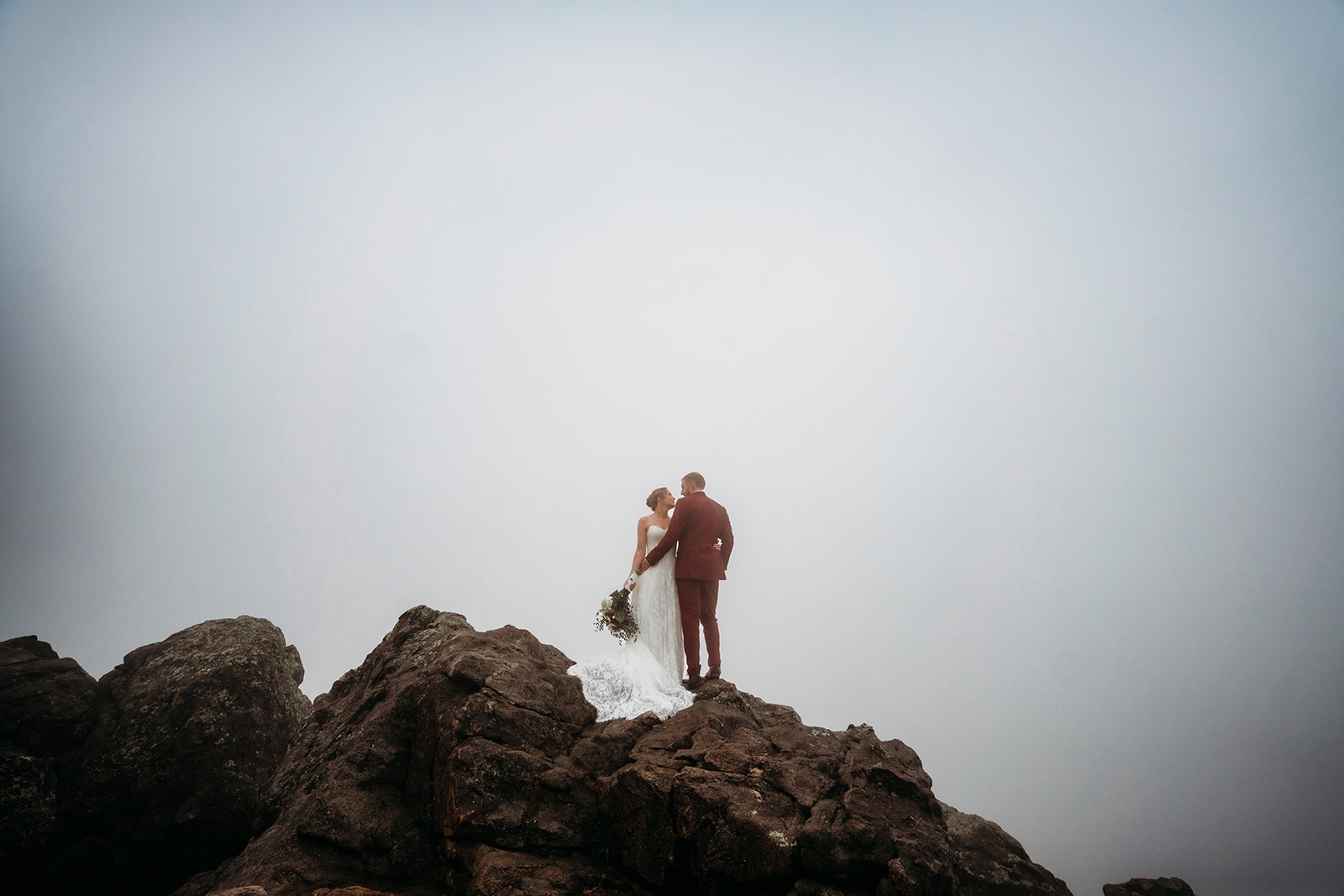 Bride and groom standing at the edge of rocky cliffs, embraced by fog, during their Colorado elopement.