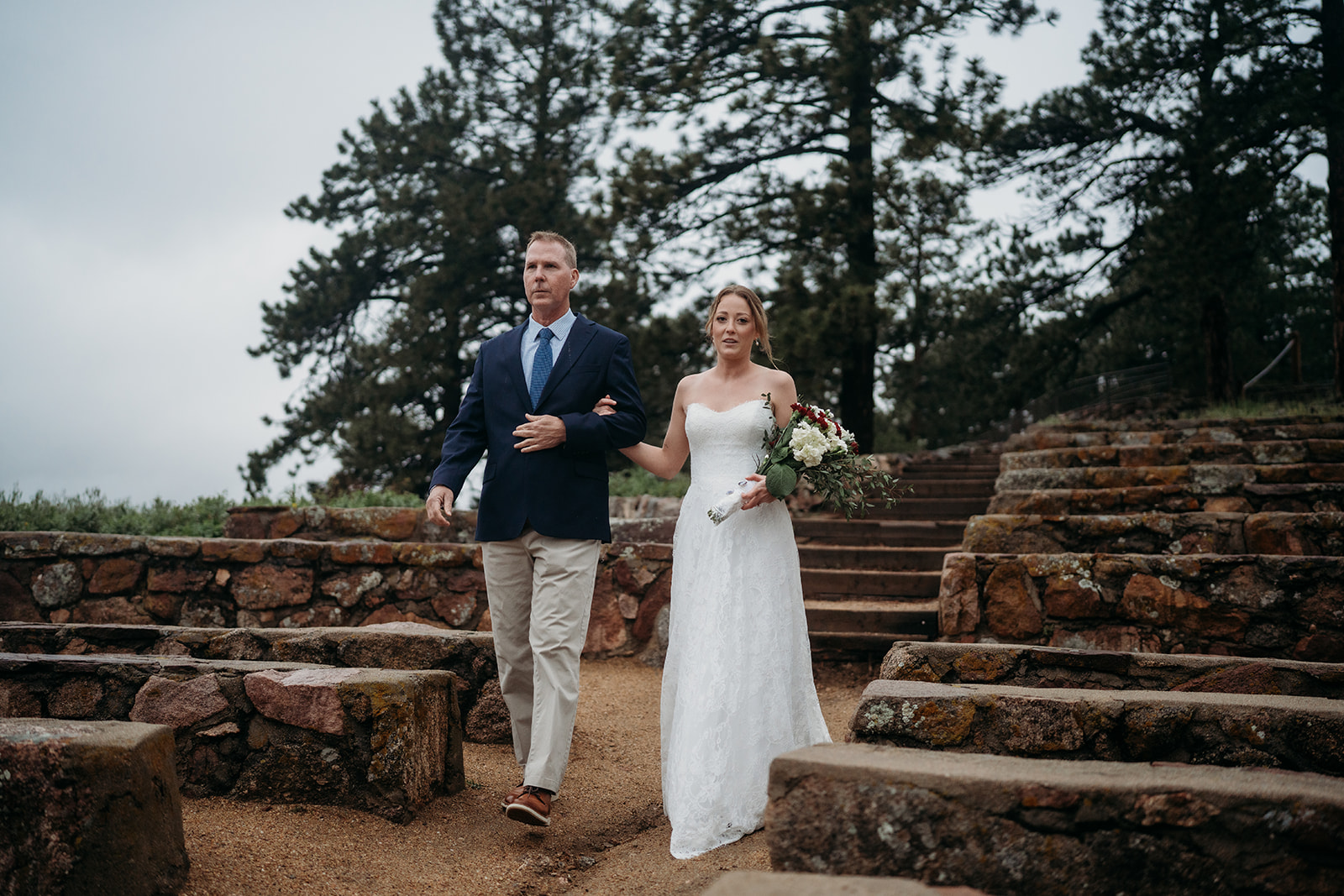 Bride and father walking together down rustic stone steps surrounded by pine trees before the ceremony.