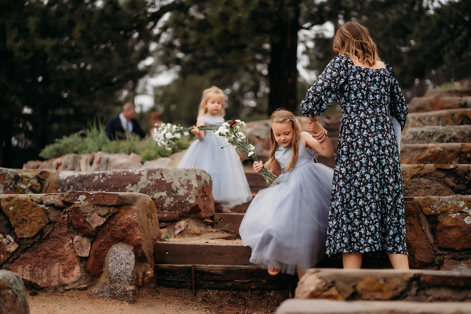 Two flower girls in blue dresses carrying flowers, carefully guided down the stone steps by a parent.