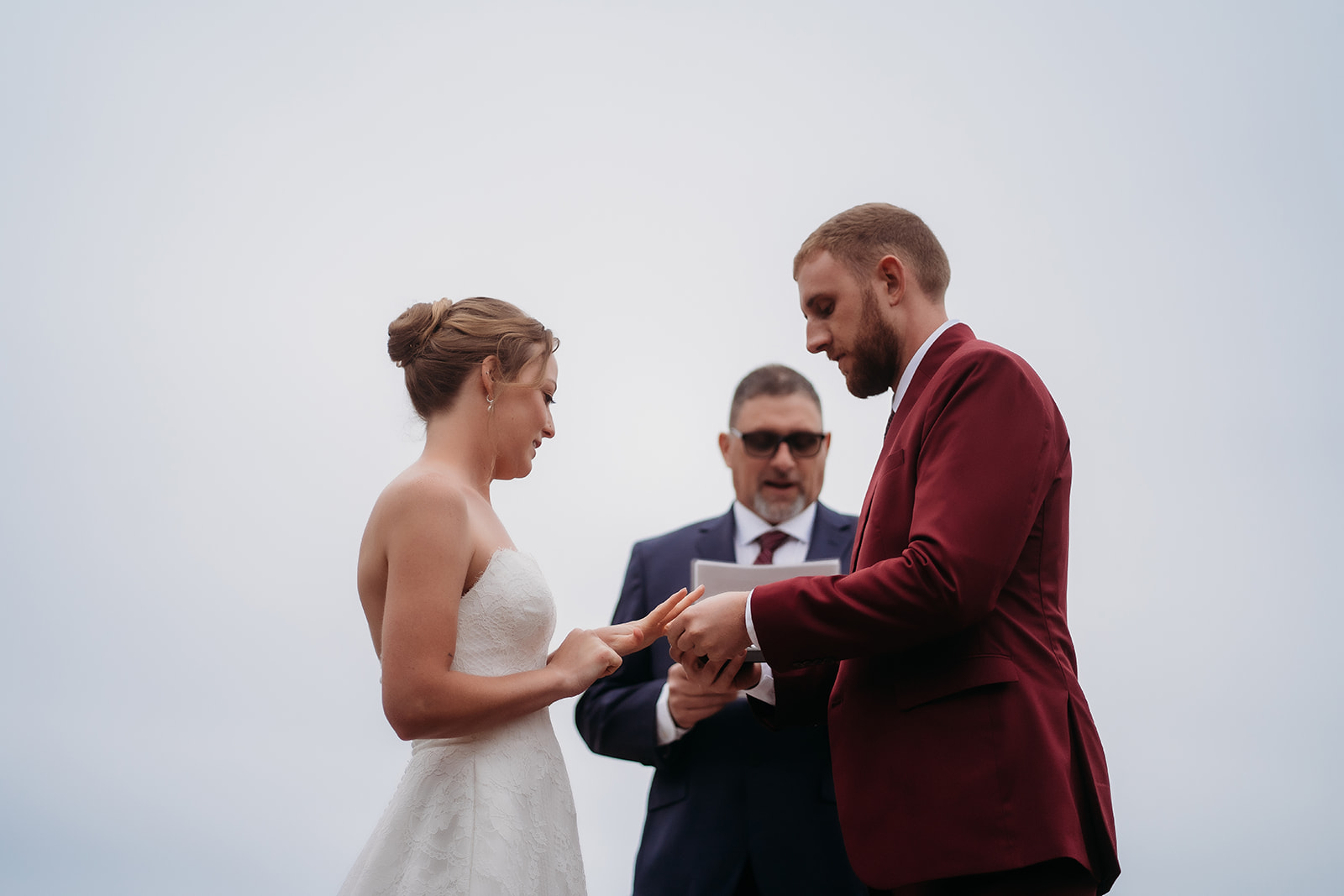 Groom places the ring on the bride’s hand while the officiant looks on.