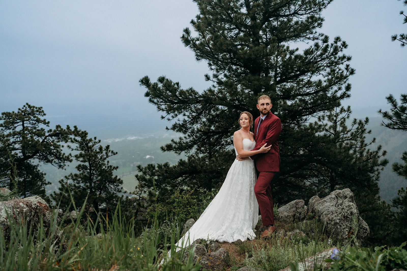Bride and groom embracing on a mountain ledge, surrounded by pine trees, a romantic example of how to plan an elopement in nature.