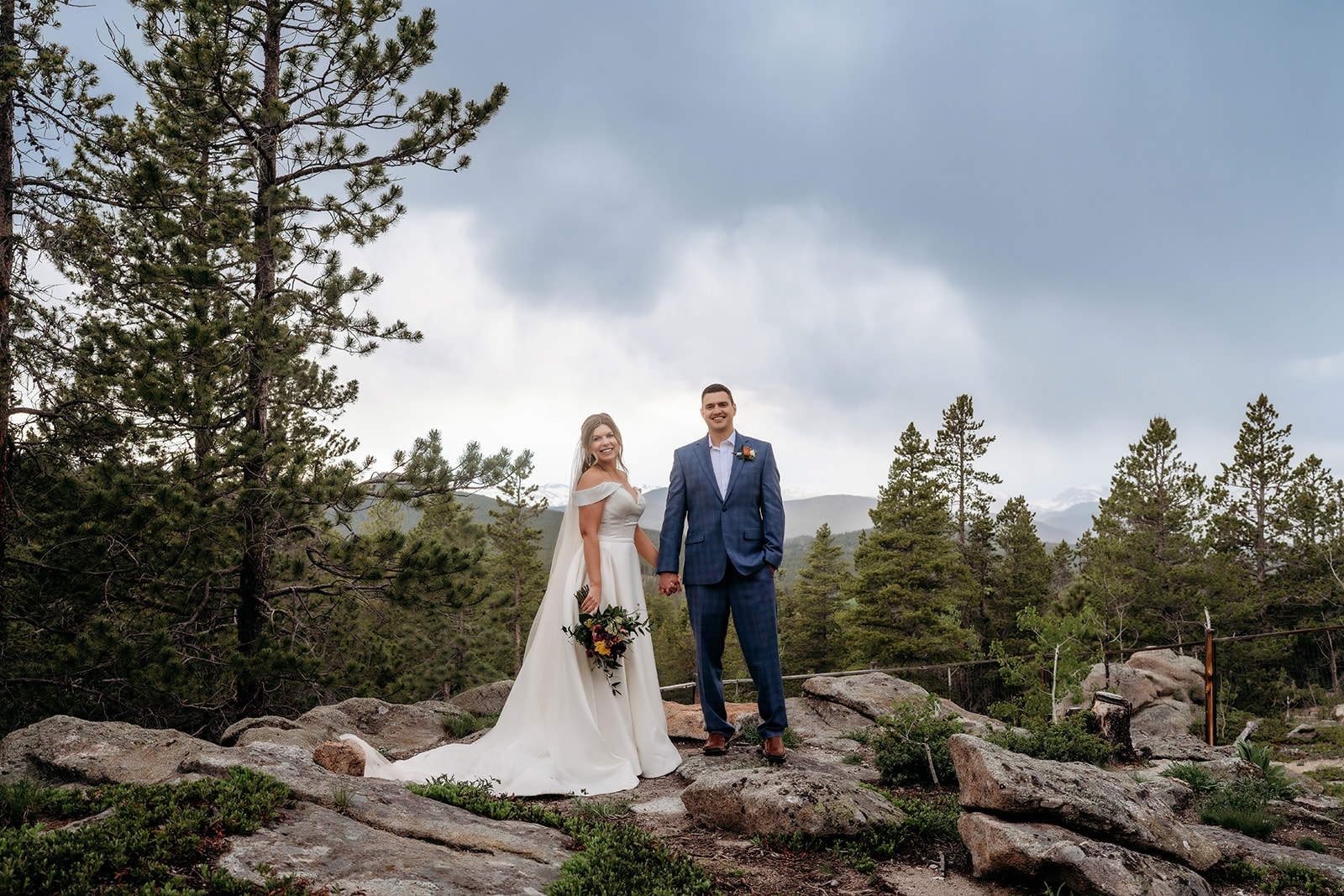 Bride and groom holding hands on rocky terrain with pine trees and mountain views at their Colorado adventure elopement.