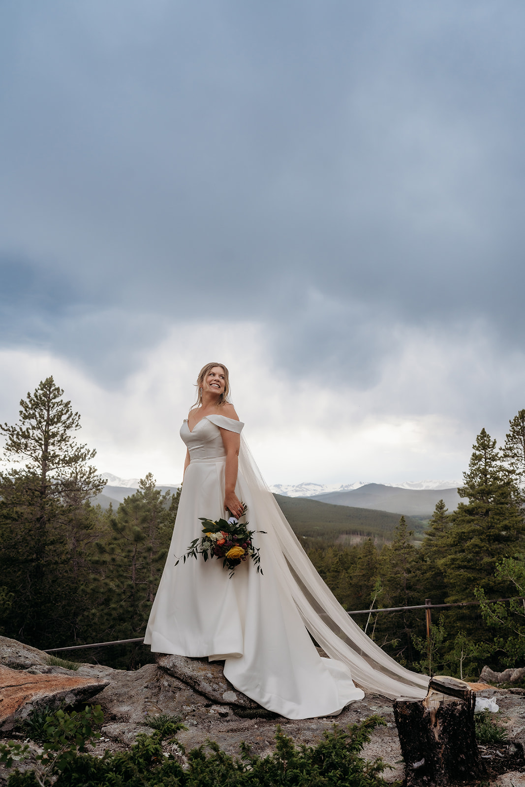 Bride in a flowing gown holding a bouquet, standing on a mountain rock with stormy skies during her Colorado adventure elopement.