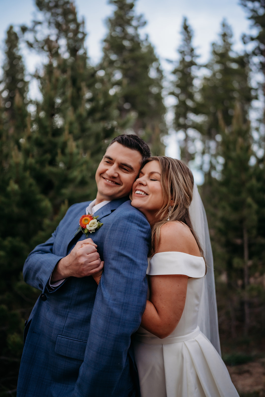 Groom in a blue suit embraces his bride in a white gown, smiling together in the forest during their Colorado adventure elopement.