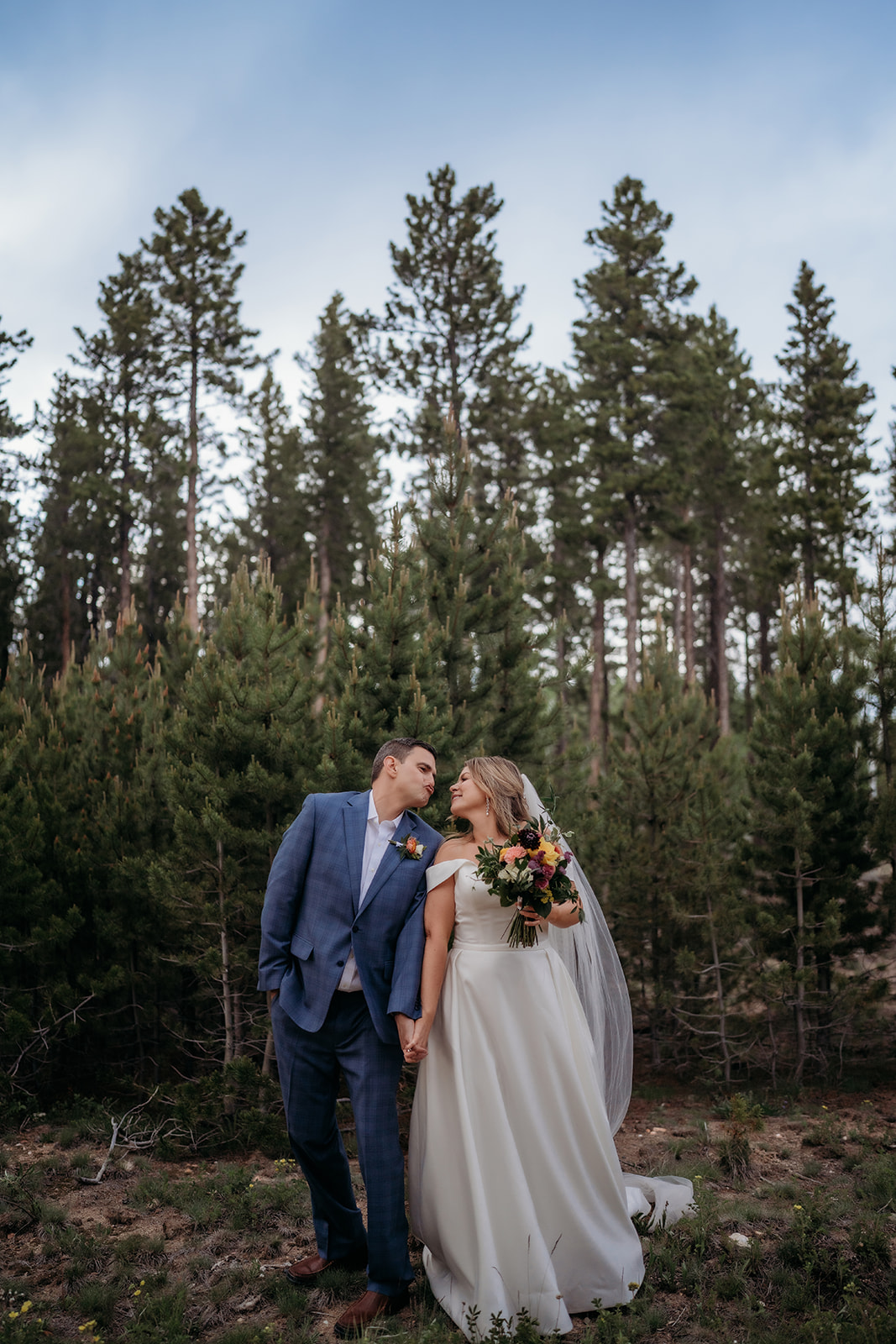 Bride and groom holding hands and leaning in for a kiss among tall pine trees at their Colorado adventure elopement.