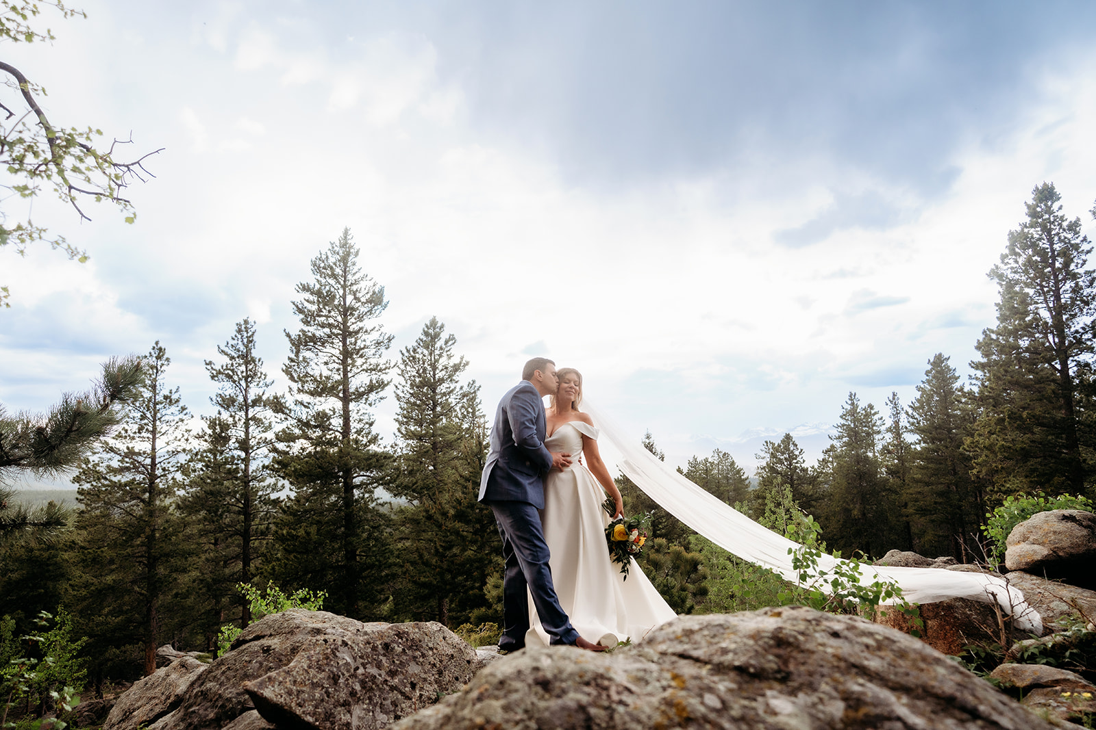 Bride and groom embracing on a rocky mountain overlook with flowing veil during their Colorado adventure elopement.