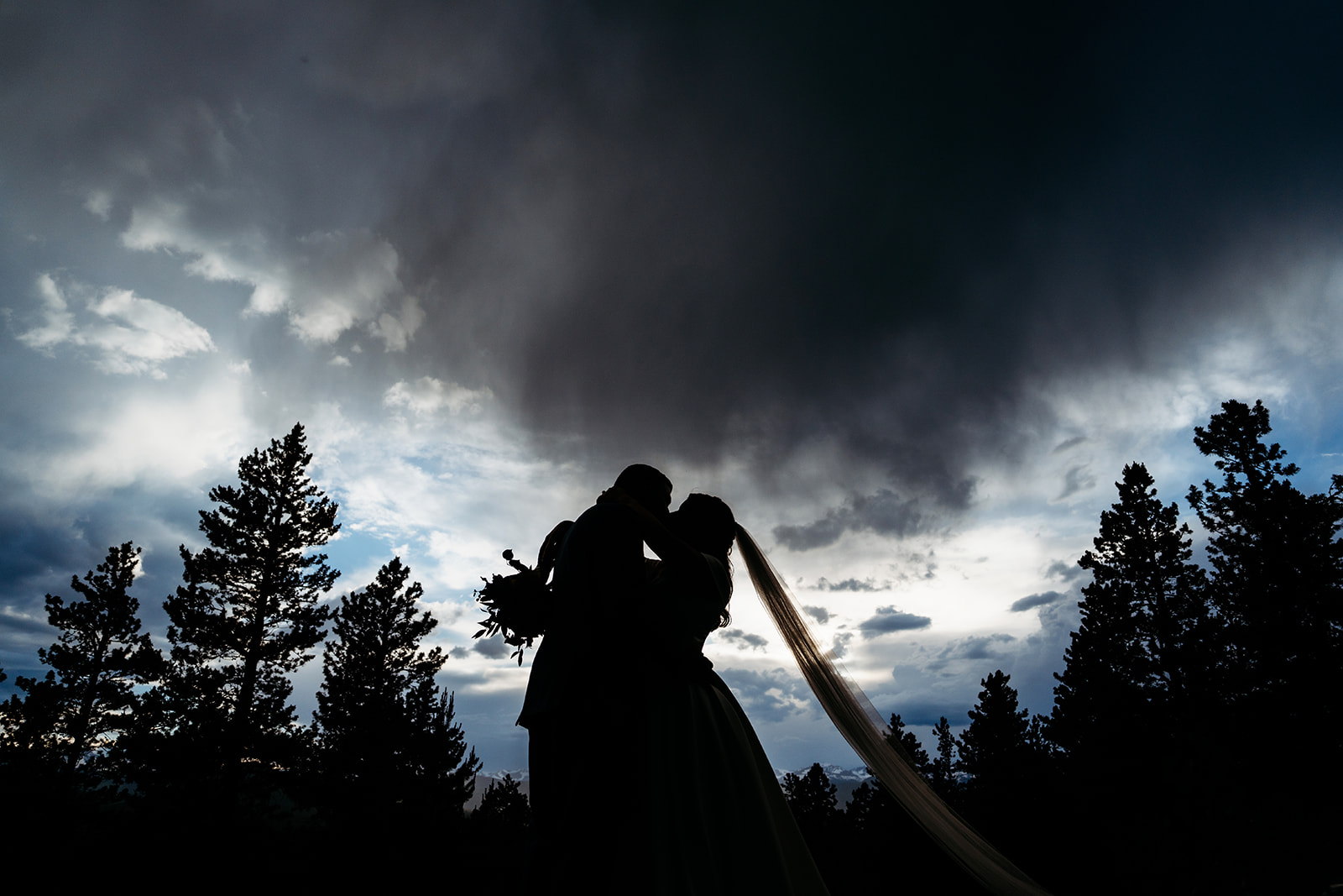 Bride and groom kiss after the sun sets on top of rock with clouds above them and bride's veil blowing in the wind