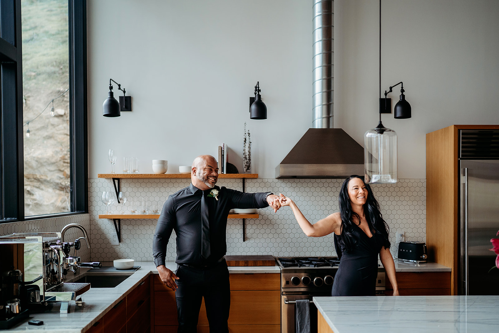 Couple laughing and holding hands while dancing in a bright kitchen with wooden accents.