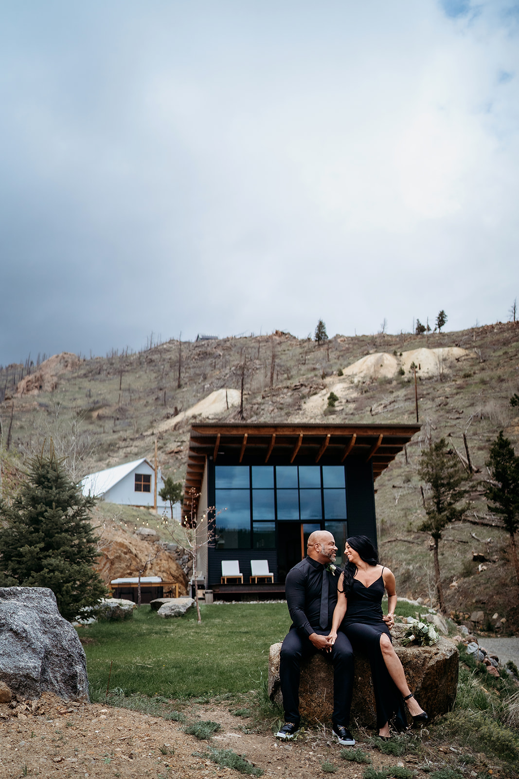 Couple sitting on a rock in front of a modern cabin in the mountains, holding hands and smiling.