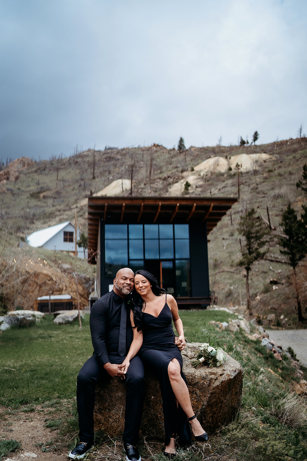 Couple sitting closely together on a rock in front of a modern cabin, sharing a quiet moment outdoors.