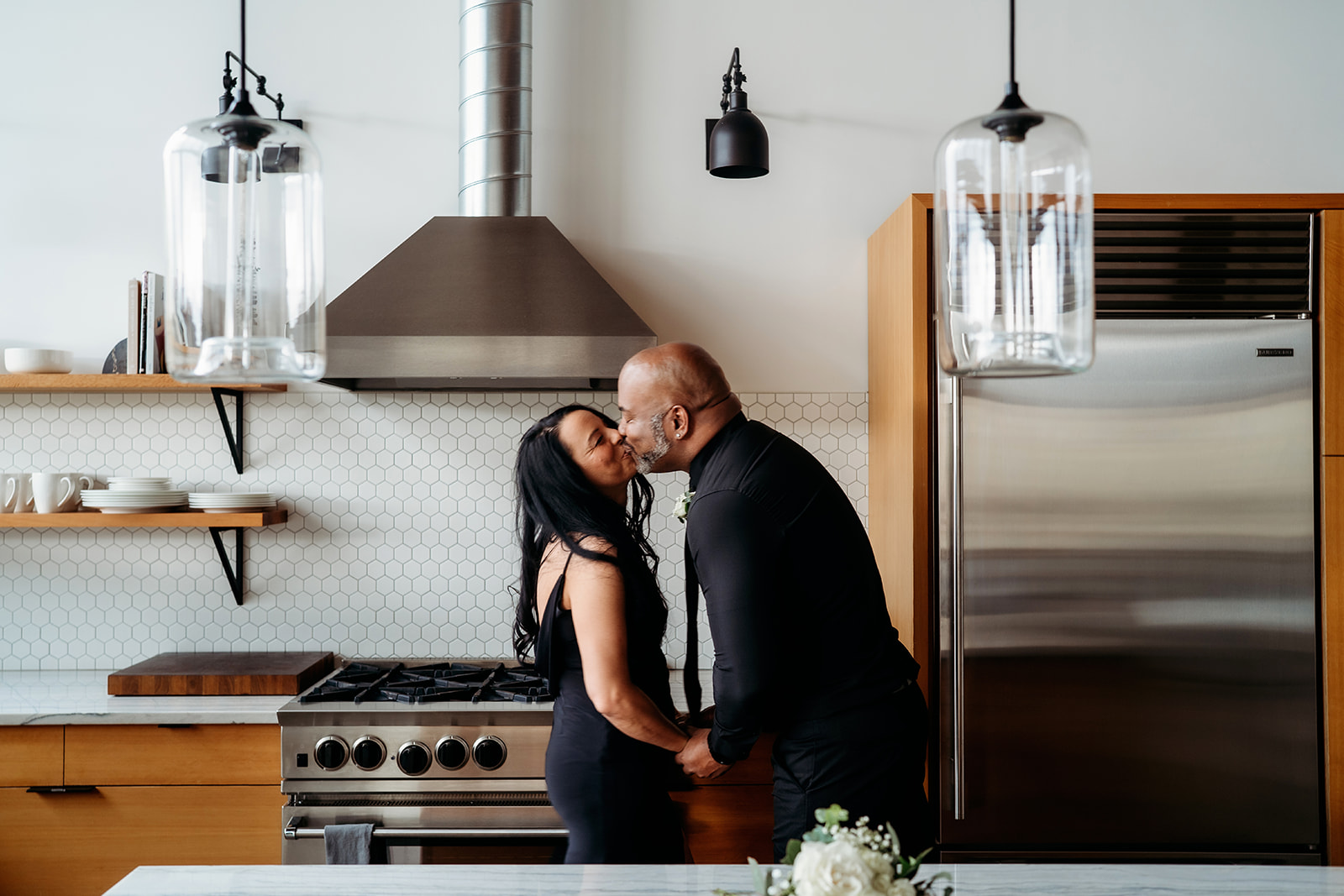 Couple kissing in a modern kitchen with white hex tile backsplash and wood cabinetry.