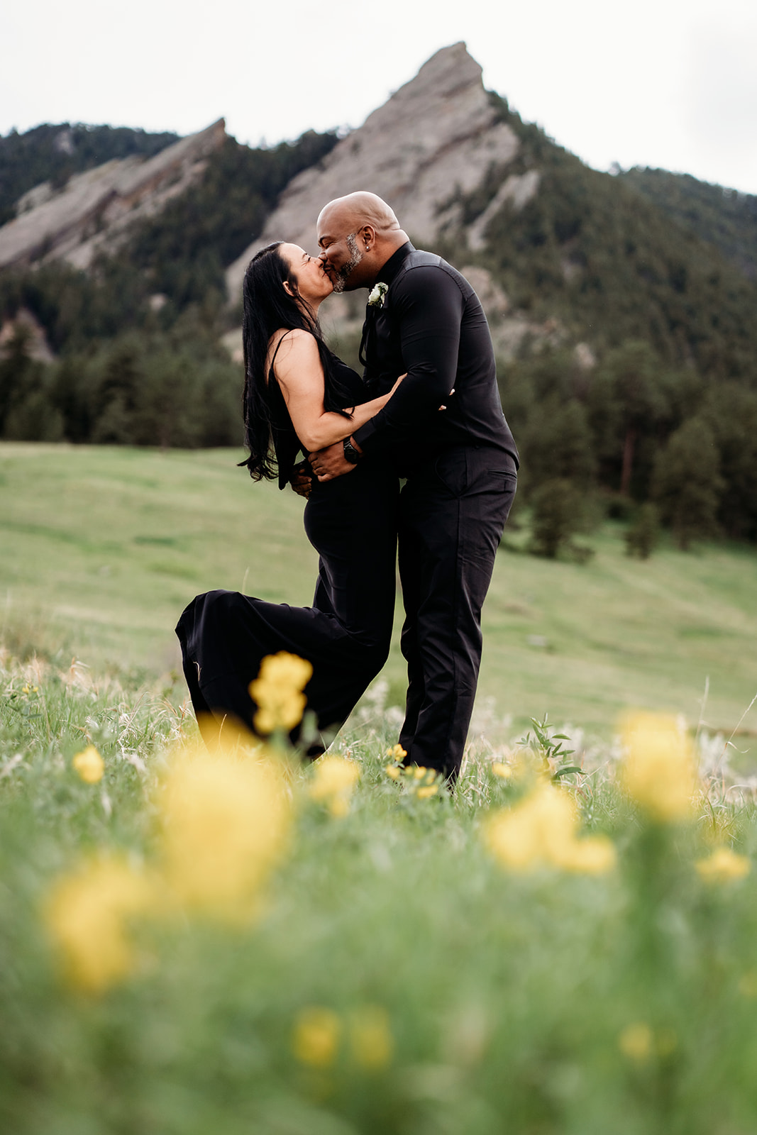 Couple sharing a kiss with wildflowers in the foreground and mountains in the background during summer engagement photos.