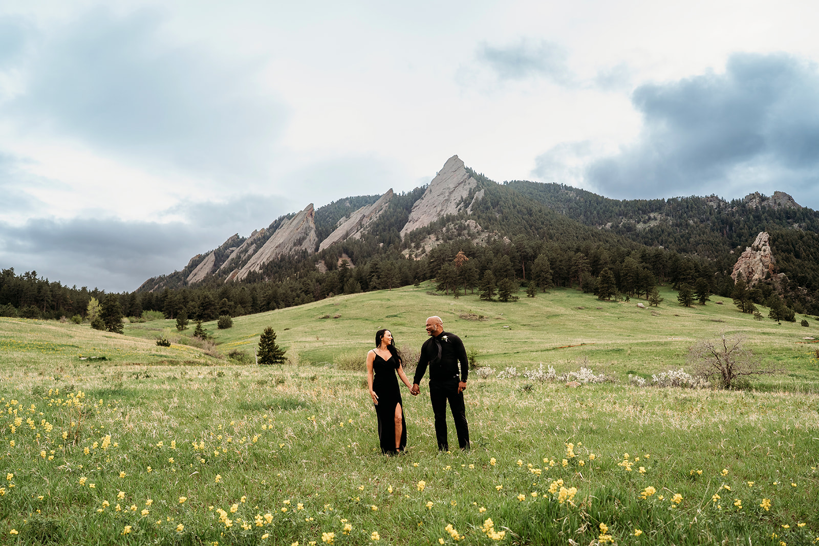 Couple holding hands and smiling at each other in a wildflower meadow with the Flatirons mountains behind during summer engagement photos.