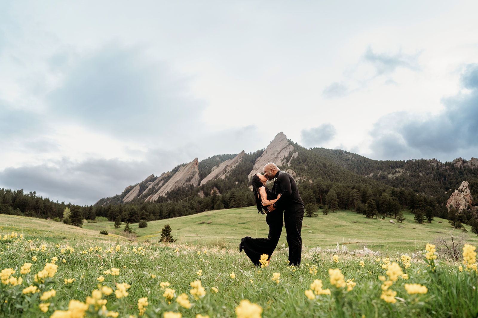 Romantic moment of a couple kissing in a yellow wildflower field with the Flatirons mountains behind them in summer engagement photos.