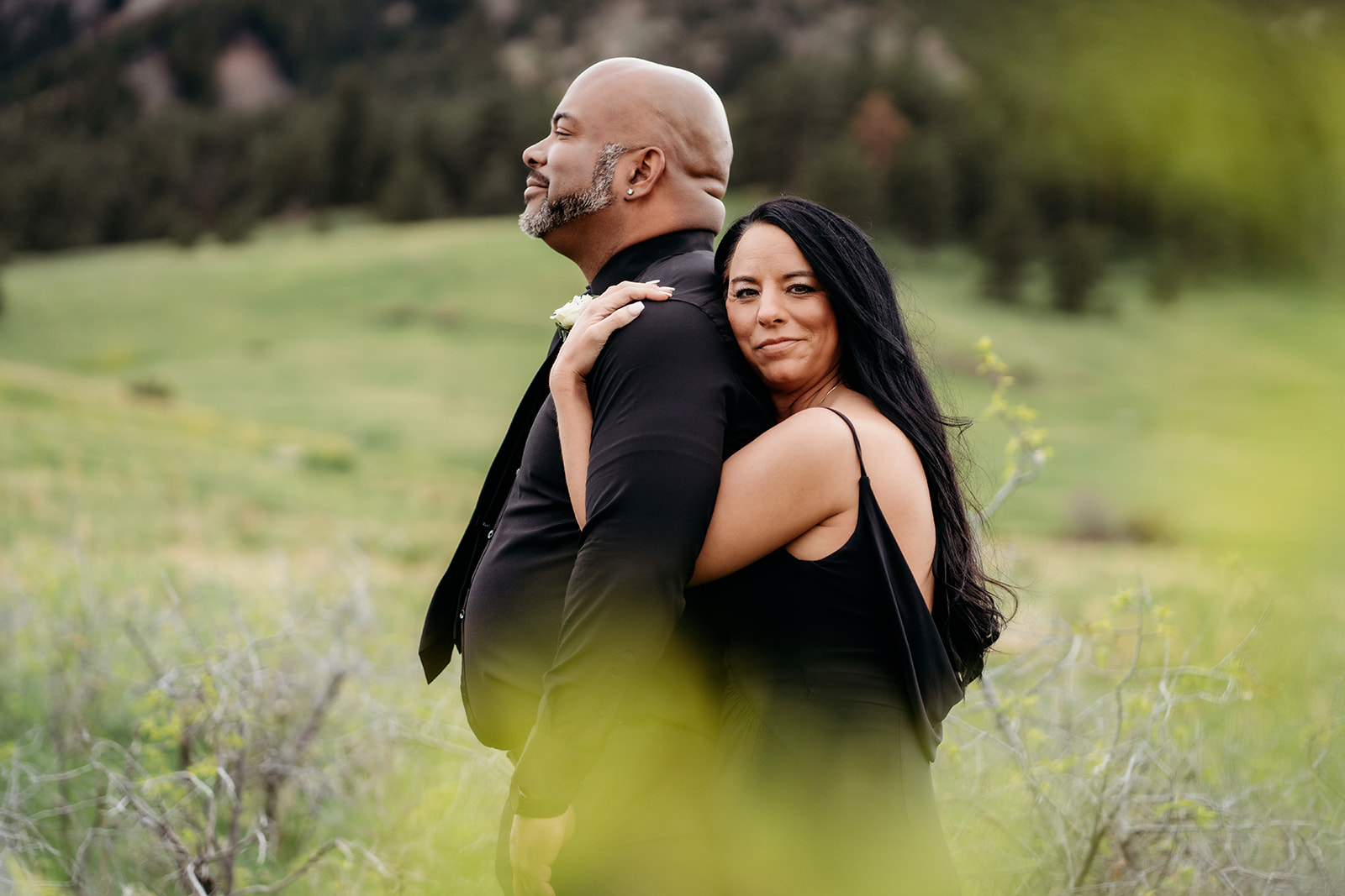Couple standing back to back in a natural setting, the woman looking confidently at the camera during summer engagement photos.