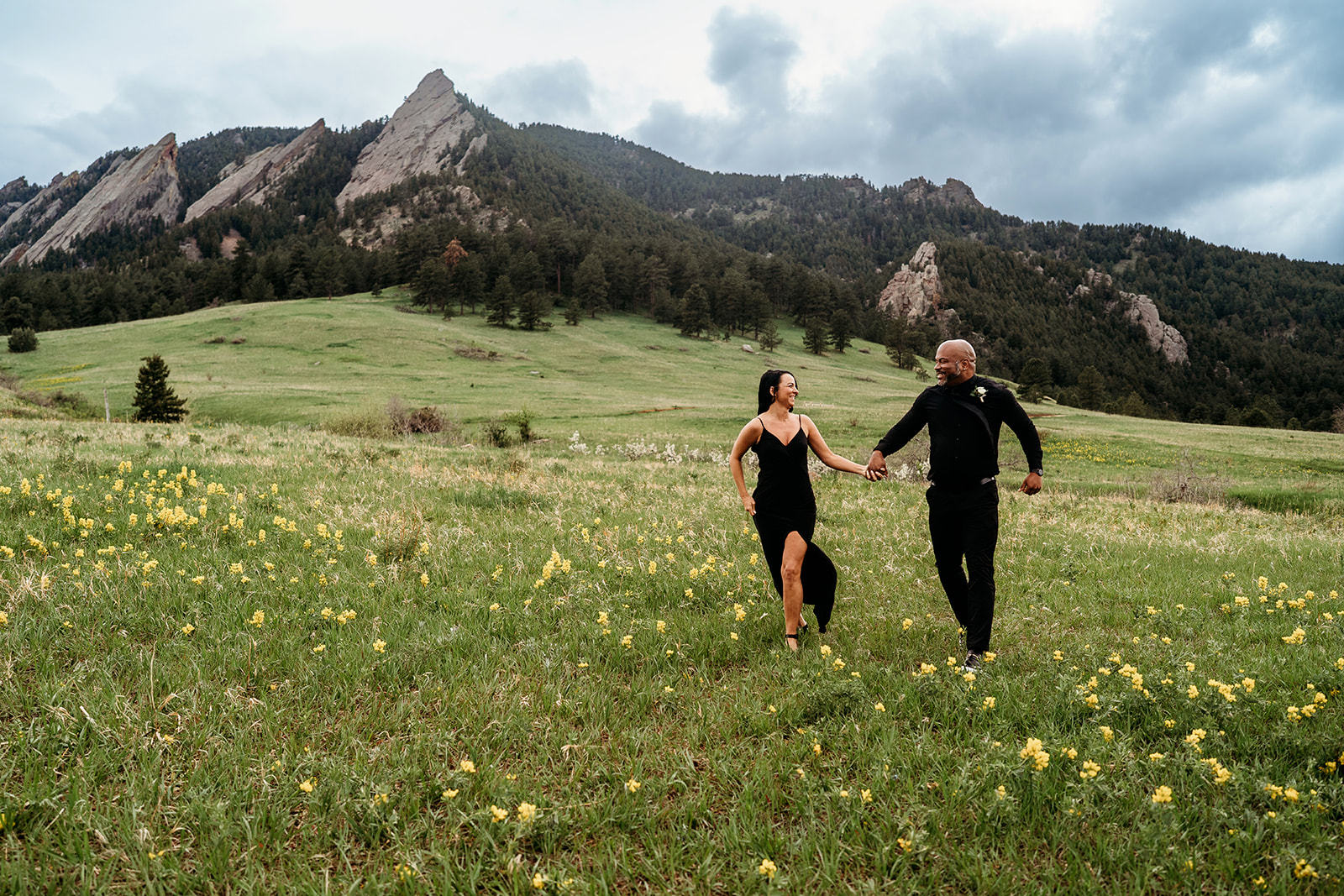 Couple holding hands and running joyfully through a wildflower-filled meadow beneath cloudy skies.