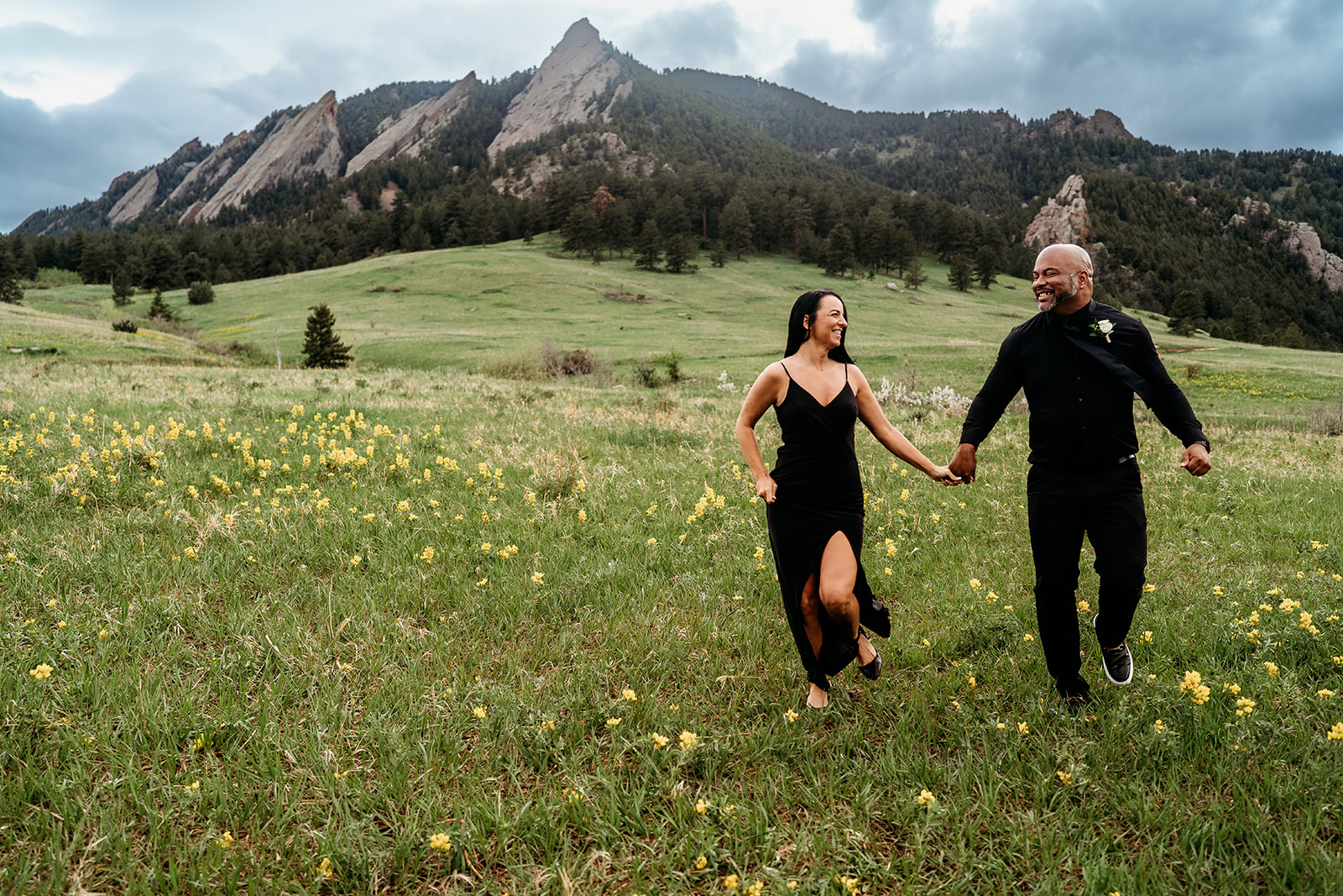Couple holding hands and running joyfully through a wildflower-filled meadow with the Flatirons mountains in the background during summer engagement photos.
