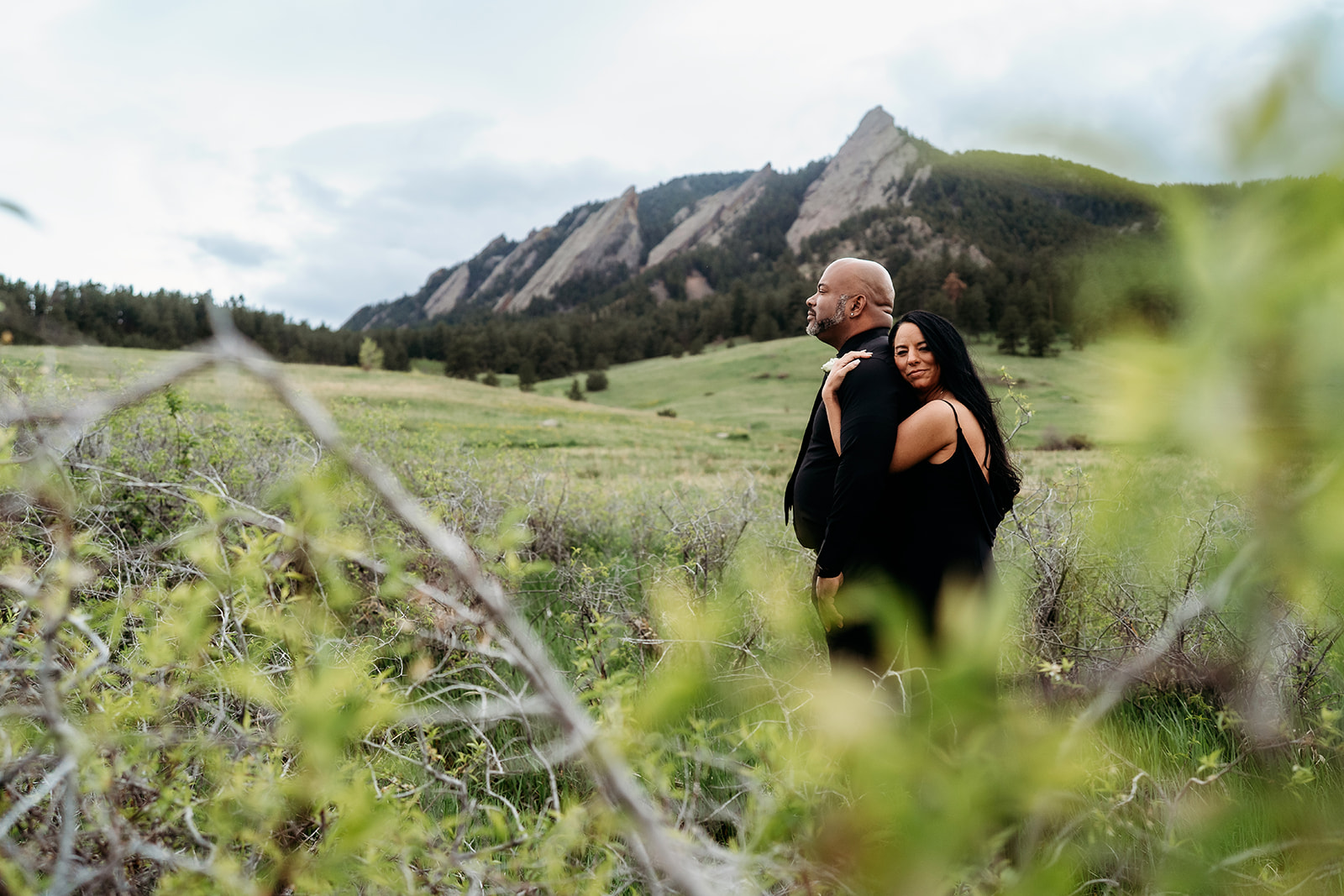 Couple embracing in a green field with the Flatirons mountains in the background during their summer engagement photoshoot.