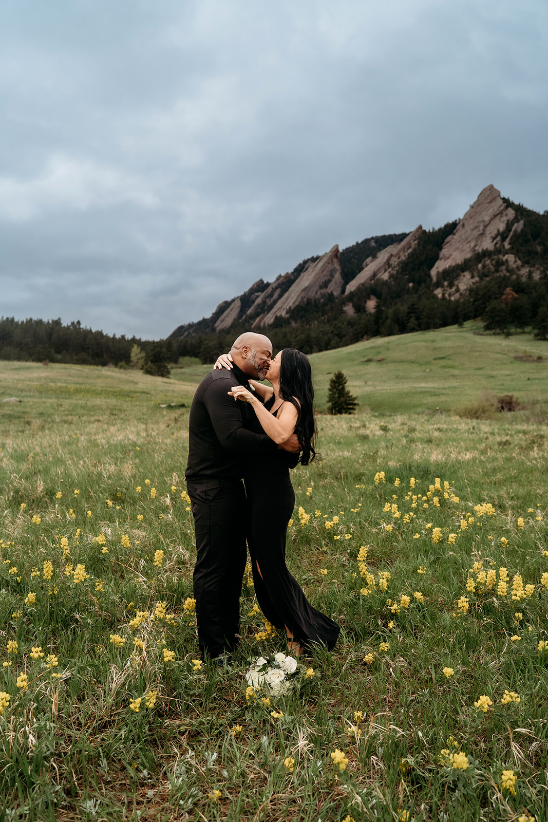 Couple embracing and smiling in a yellow wildflower field with the Flatirons mountains behind them in summer engagement photos.