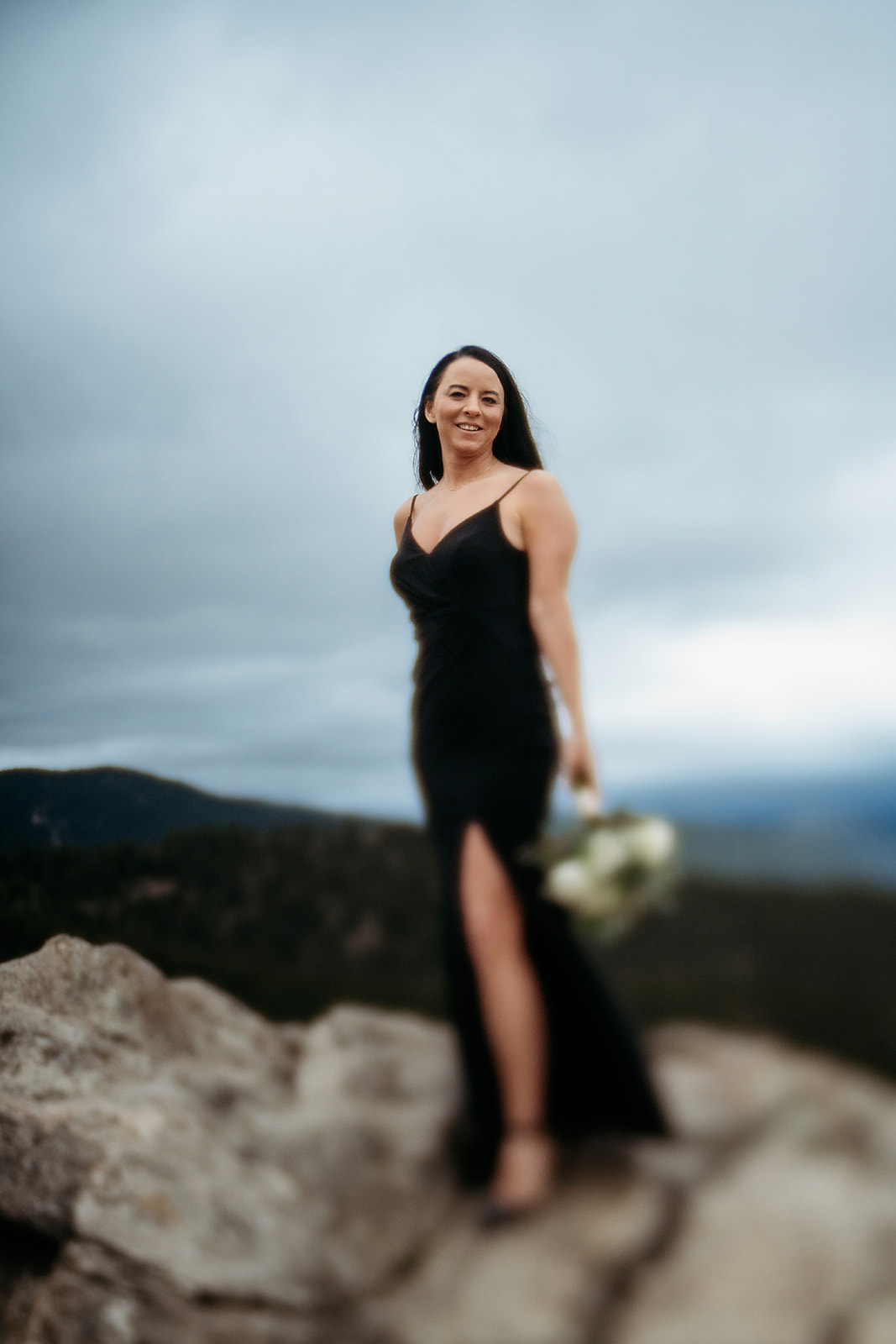 Woman in a black dress holding a bouquet standing on a rocky overlook with soft focus mountain background.