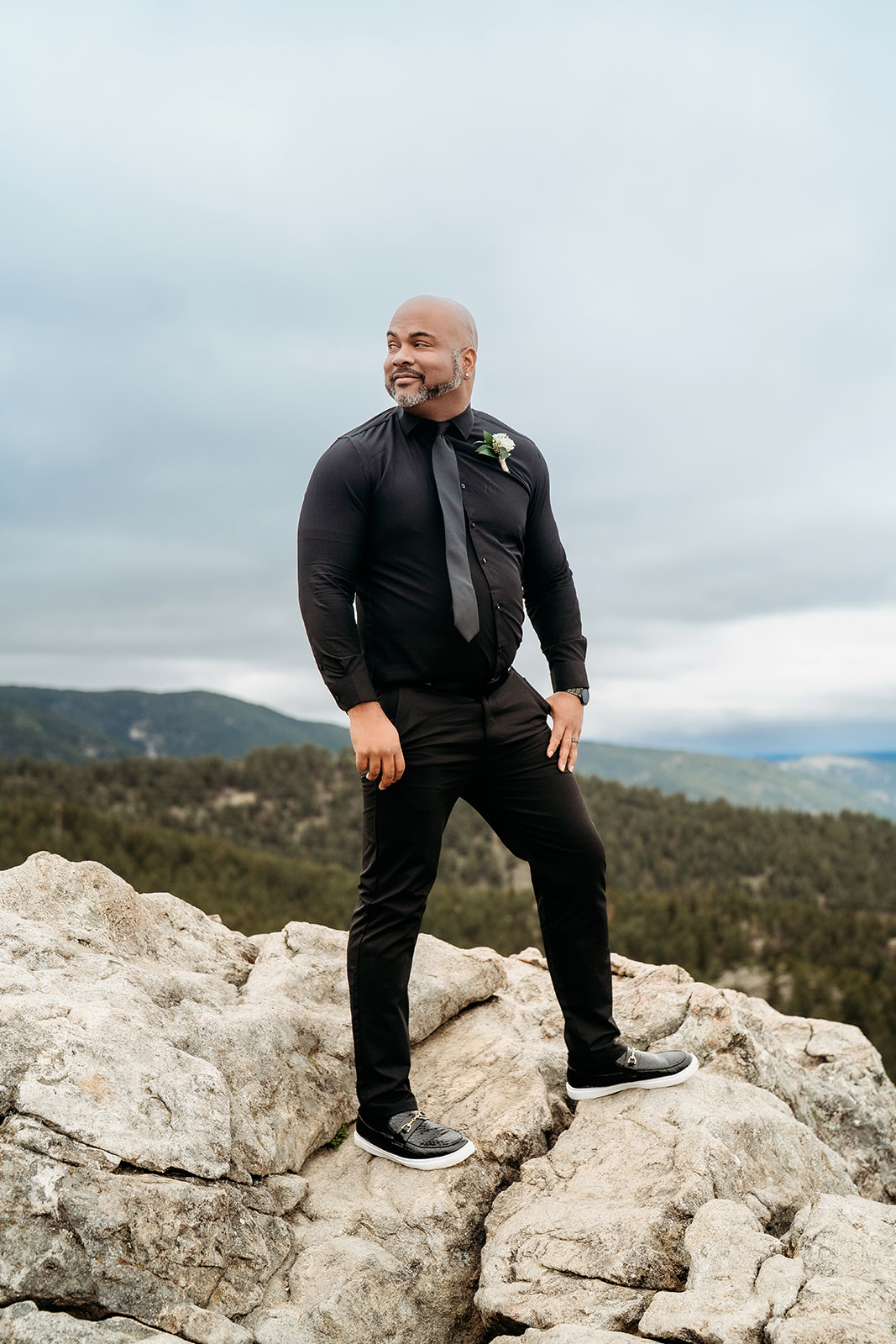 Man in black dress clothes standing confidently on rocky terrain with forested hills behind.