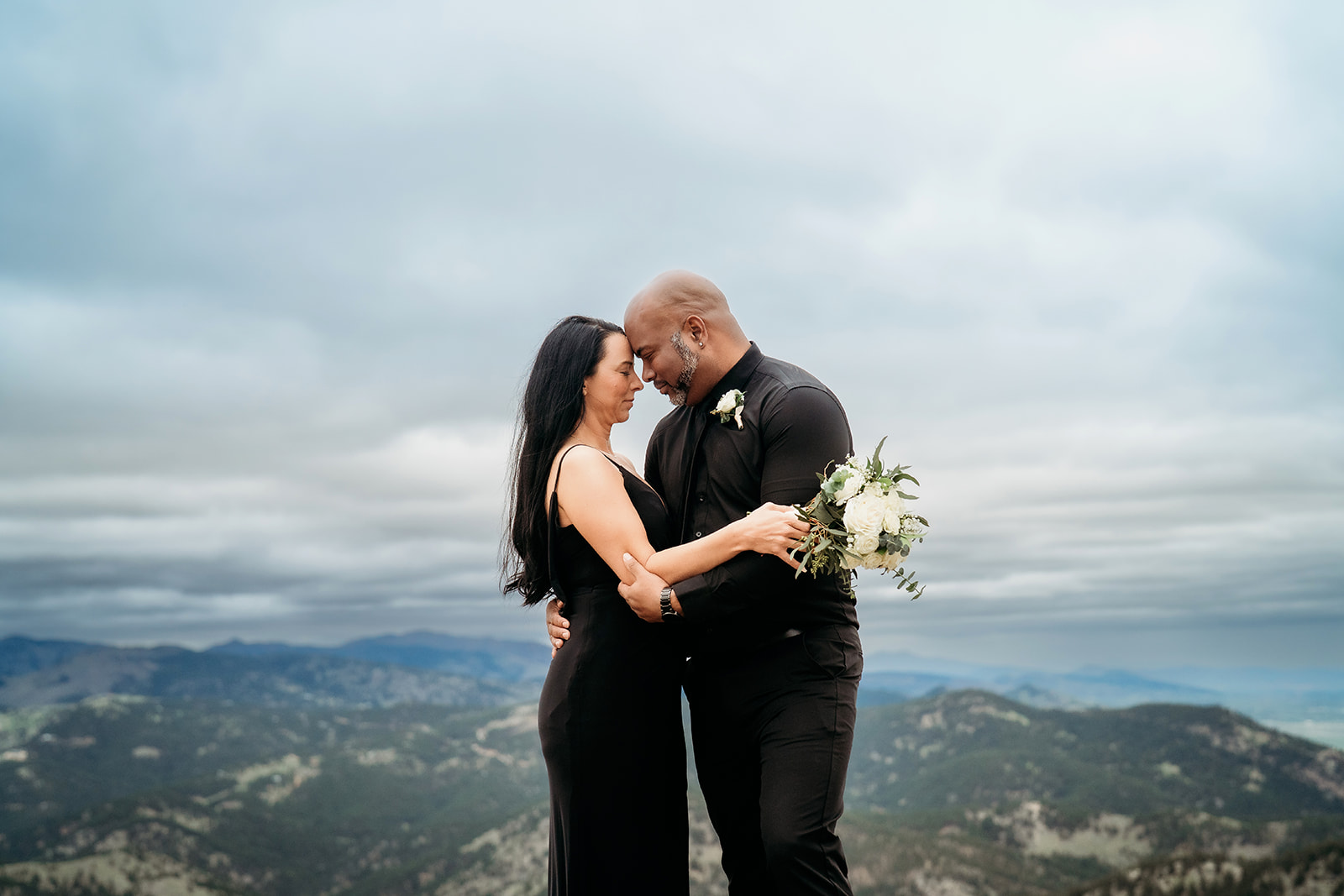 Couple standing forehead to forehead holding a bouquet of white flowers with mountain views behind them during summer engagement photos