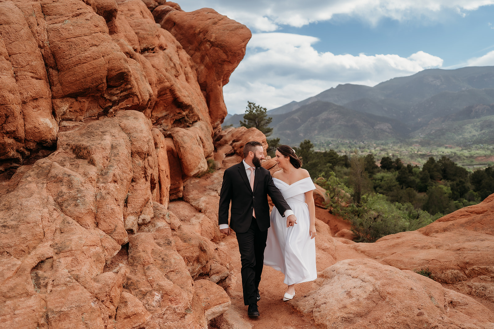 Bride and groom walking hand in hand through red rock formations at Garden of the Gods, with dramatic mountain backdrop.