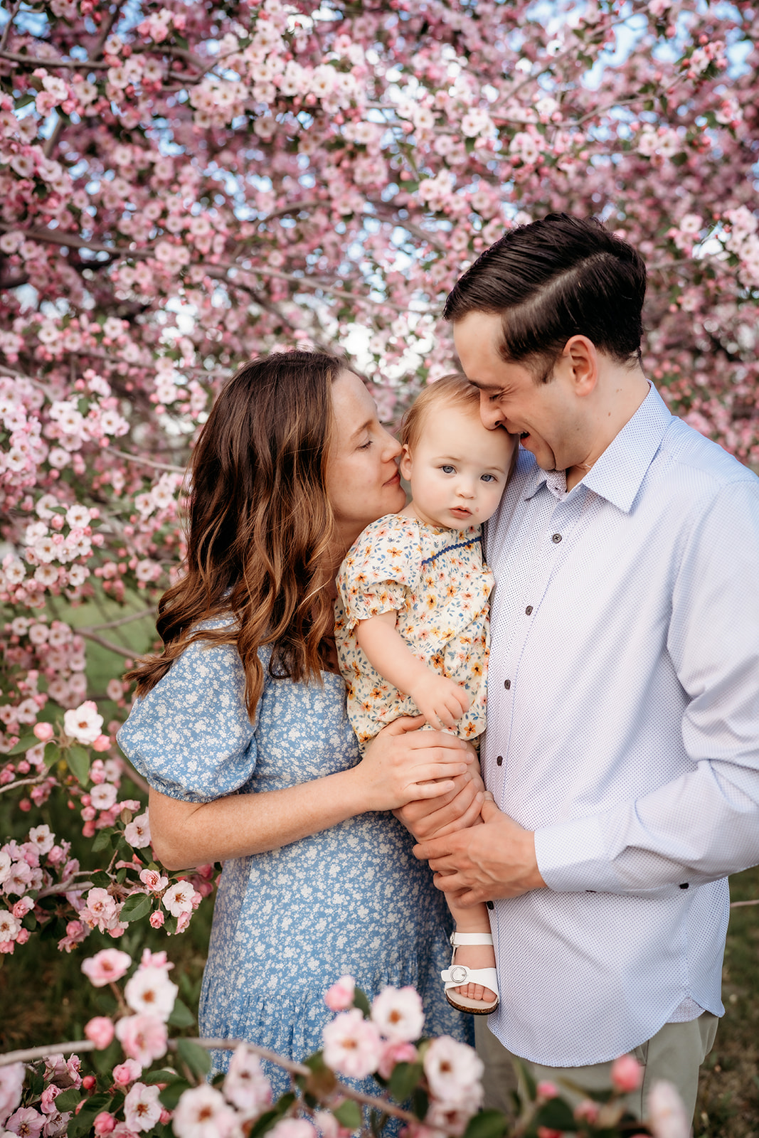 Parents holding their baby under blooming pink flowers, showing spring outfits for family photos.