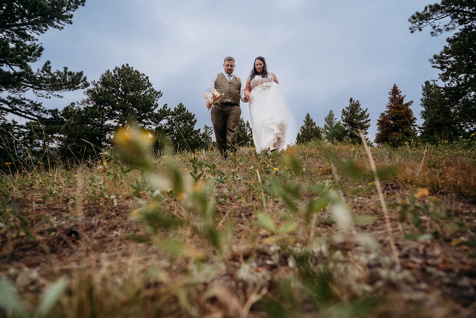 Bride and groom walk hand-in-hand down a hillside trail surrounded by wild grasses and towering pines during their intimate wedding.