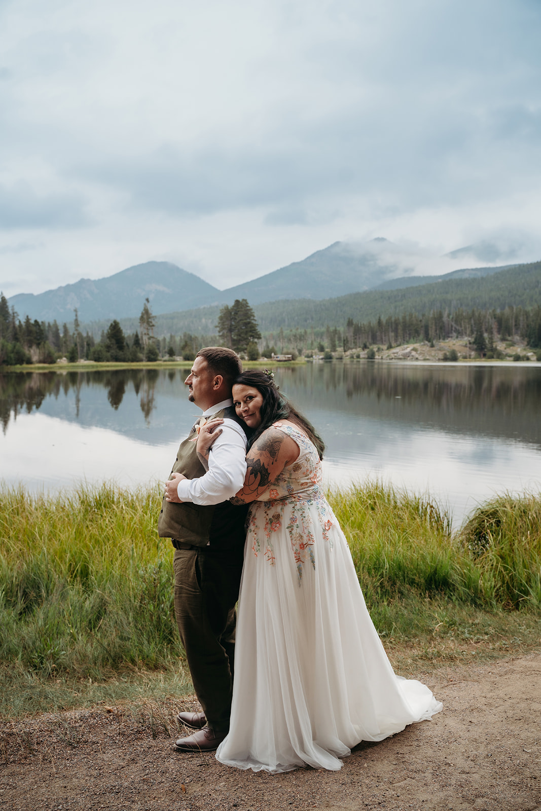 Bride wraps her arms around the groom from behind, standing together by a serene lake during their intimate wedding.