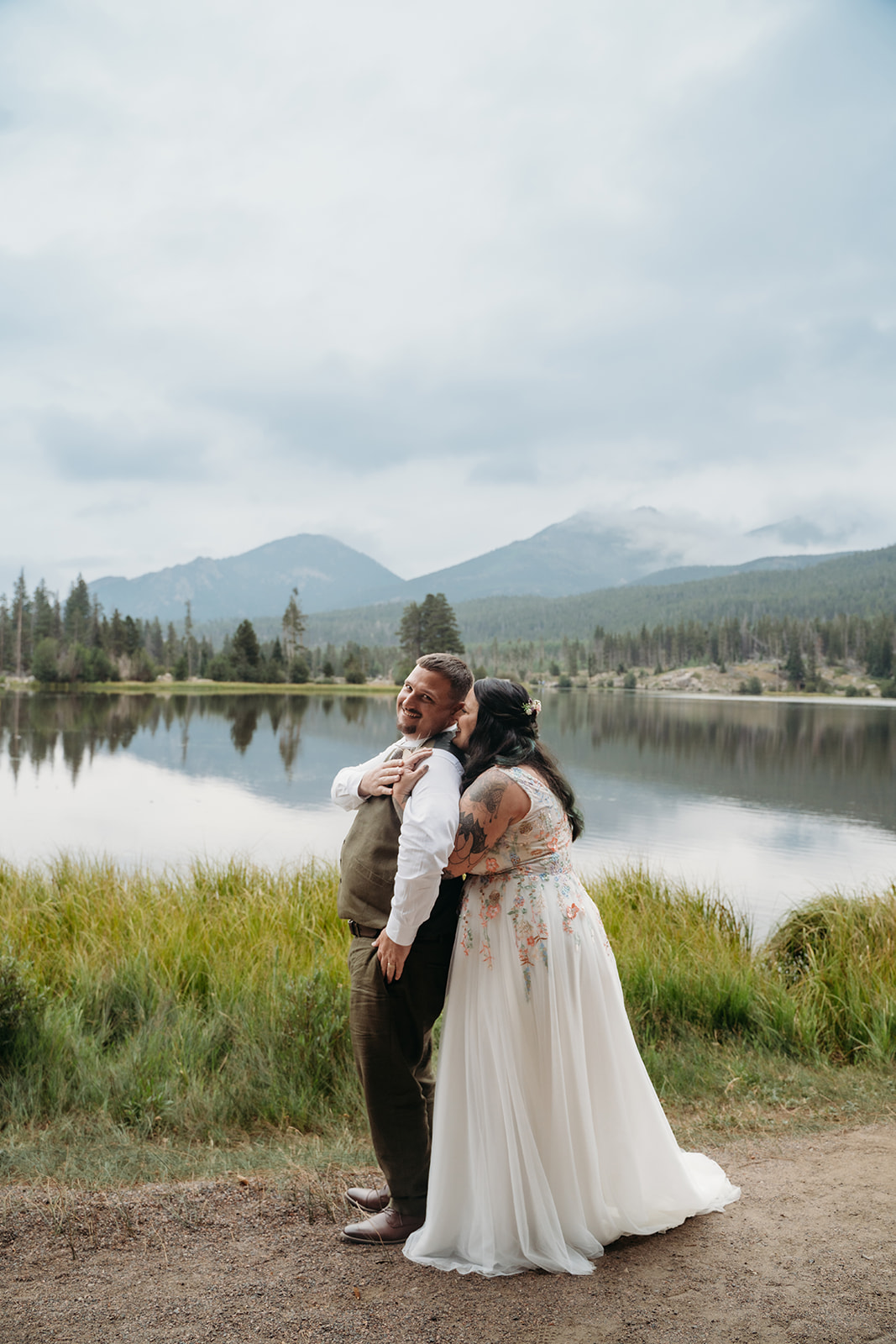Bride hugs groom from behind as they take in the mountain views along the lakeshore, sharing a peaceful moment together.