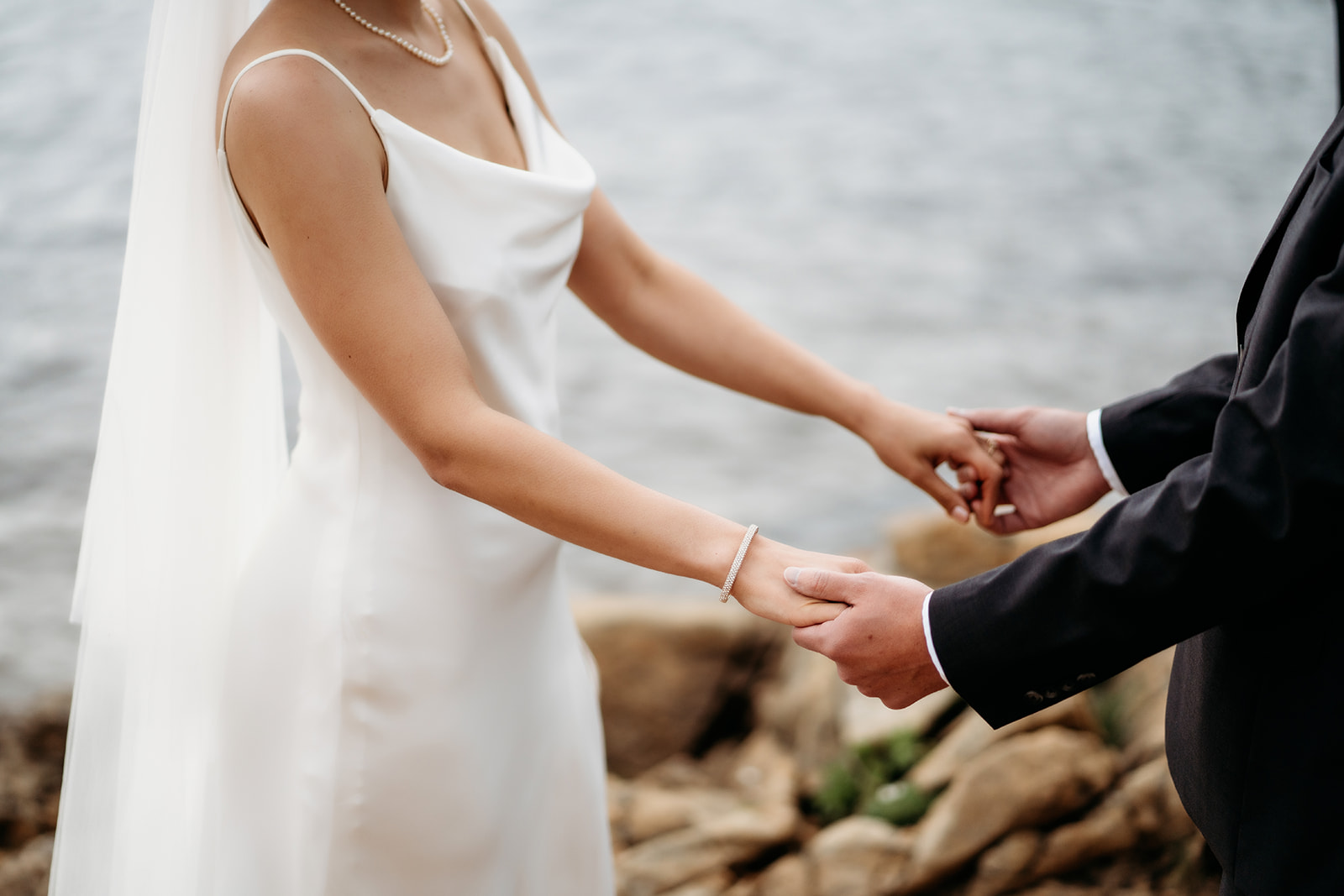 Close-up of bride and groom holding hands beside a rocky shoreline during their elopement.