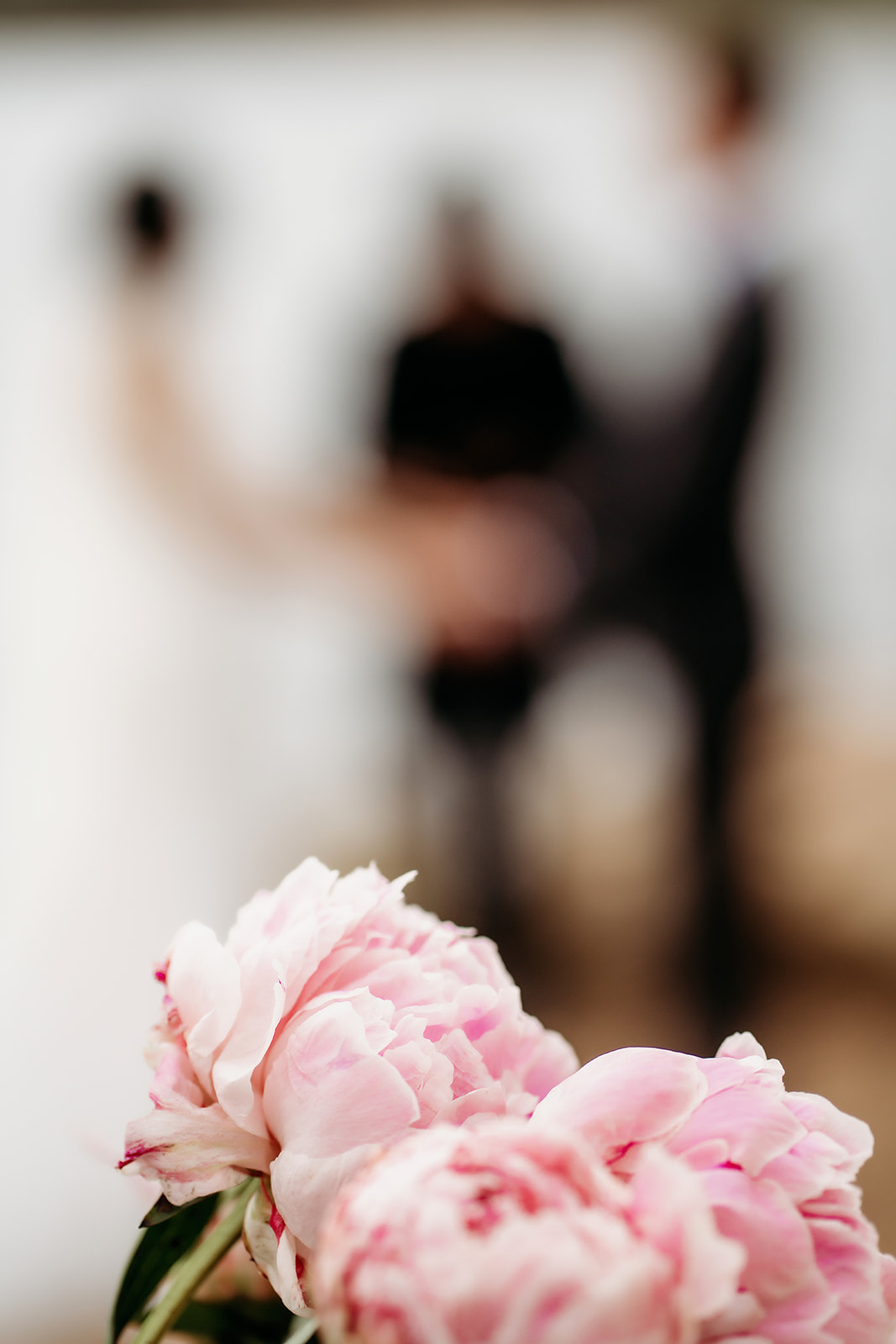 Soft pink peonies in the foreground with the couple and officiant blurred in the background, capturing romantic wedding details.
