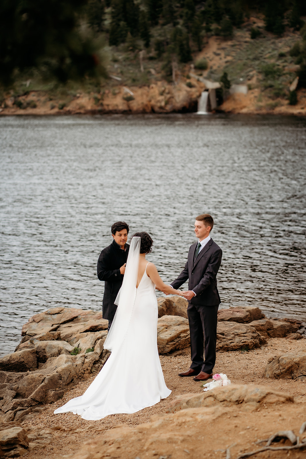 Wide shot of a couple holding hands during their lakeside ceremony with a cascading waterfall in the background, highlighting a hidden gem among Colorado elopement locations.