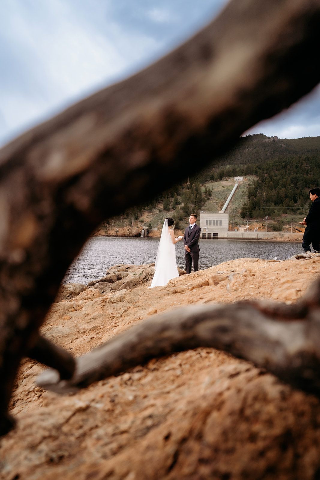 Framed through a twisted tree branch, the couple exchanges vows with a mountain reservoir as their ceremony backdrop—one of many scenic Colorado elopement locations.