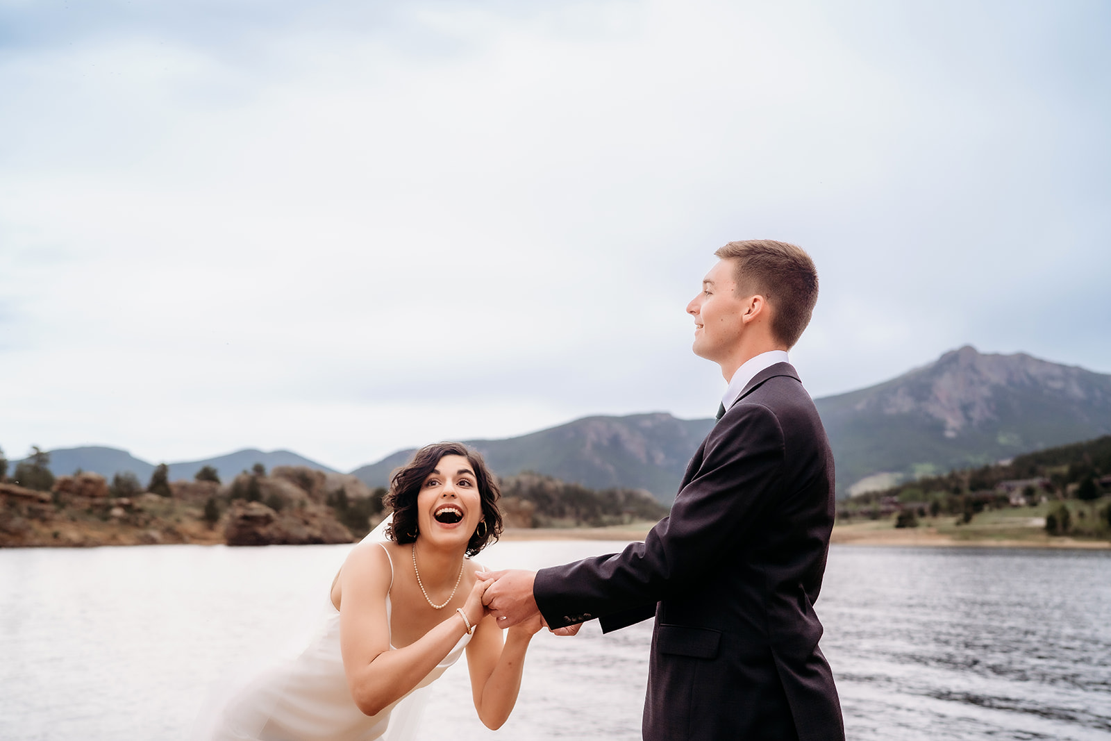 Playful moment between bride and groom as they laugh beside the water with mountain peaks rising behind them.